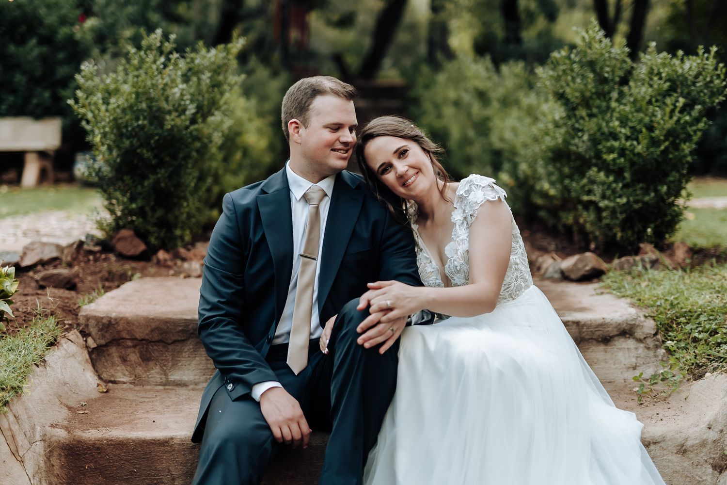 Happy newlyweds sit together on stone steps in a garden, bride in white gown and groom in navy suit.