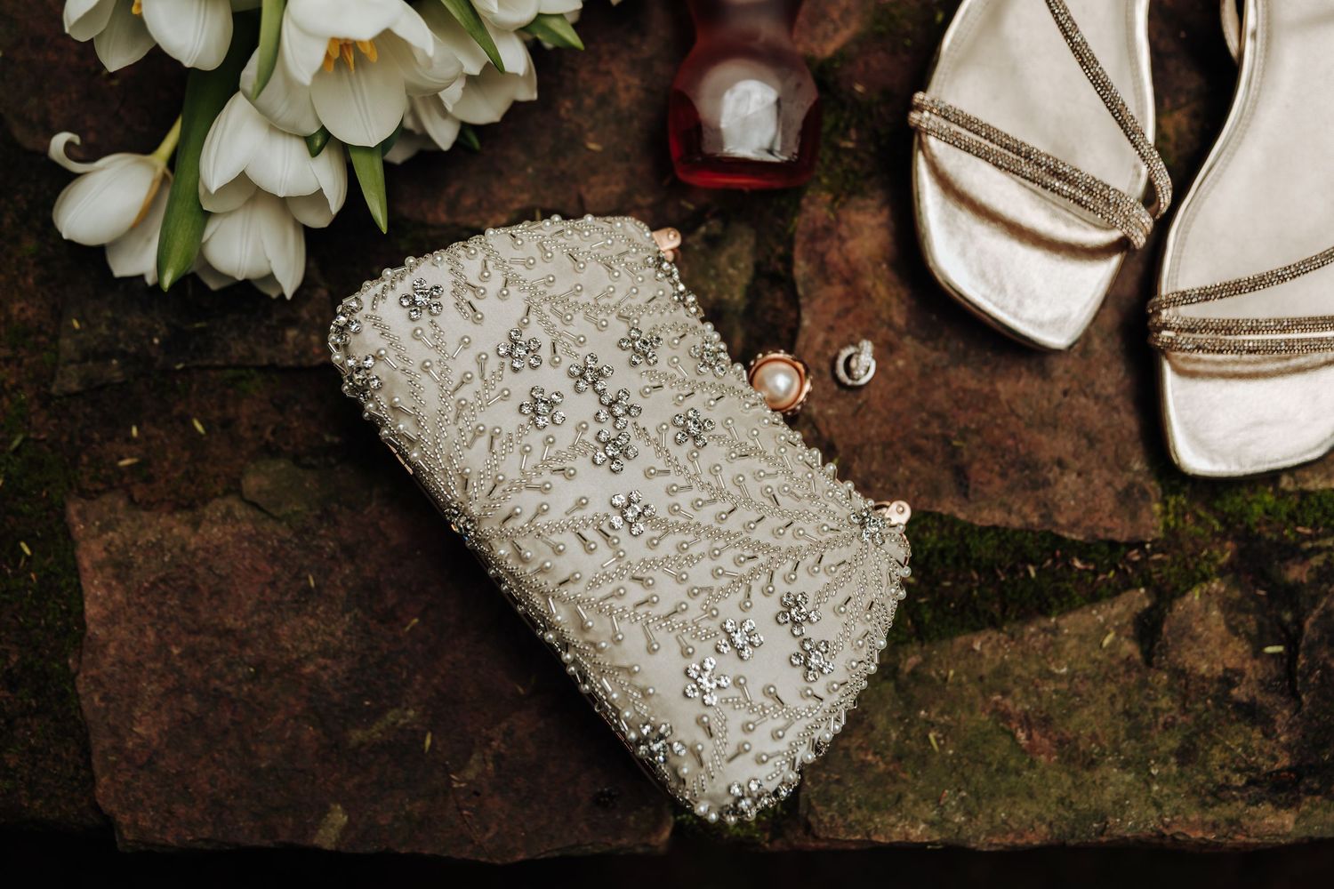 Elegant beaded white bridal clutch purse displayed on stone with white flowers, gold heels, and jewelry accessories.