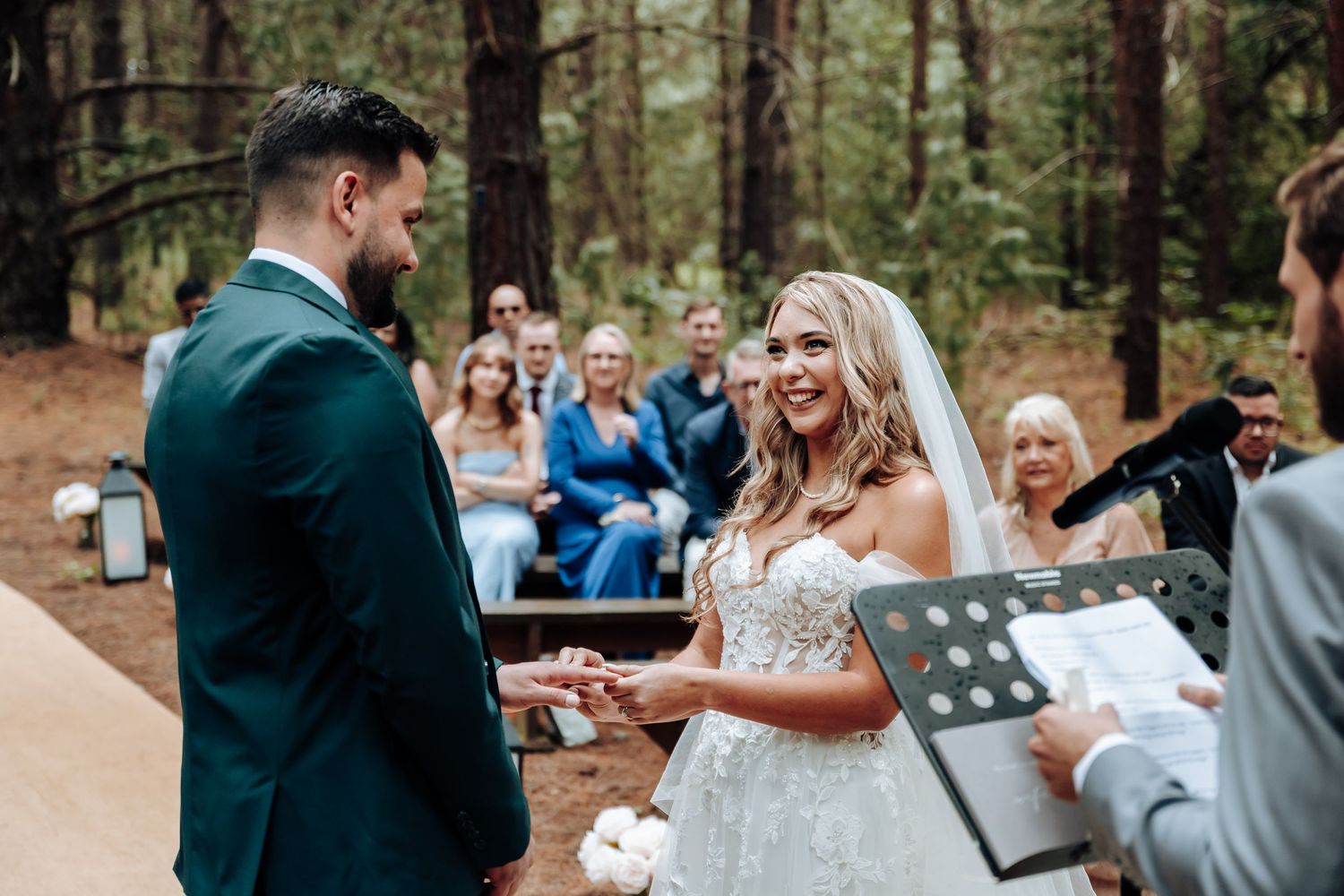 A couple exchanges rings during an outdoor forest wedding ceremony surrounded by guests and tall pine trees.