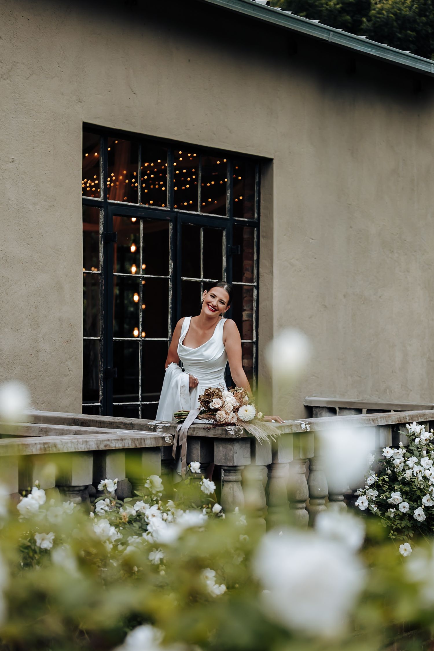 A person in white sits on a rustic porch with string lights and white flowers in the garden foreground.