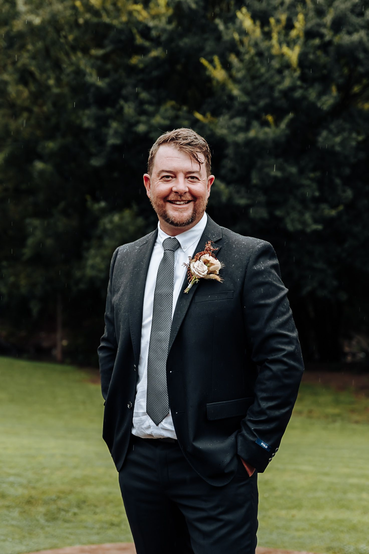 A groom in a black suit and white tie poses outdoors in a grassy area with trees in the background.