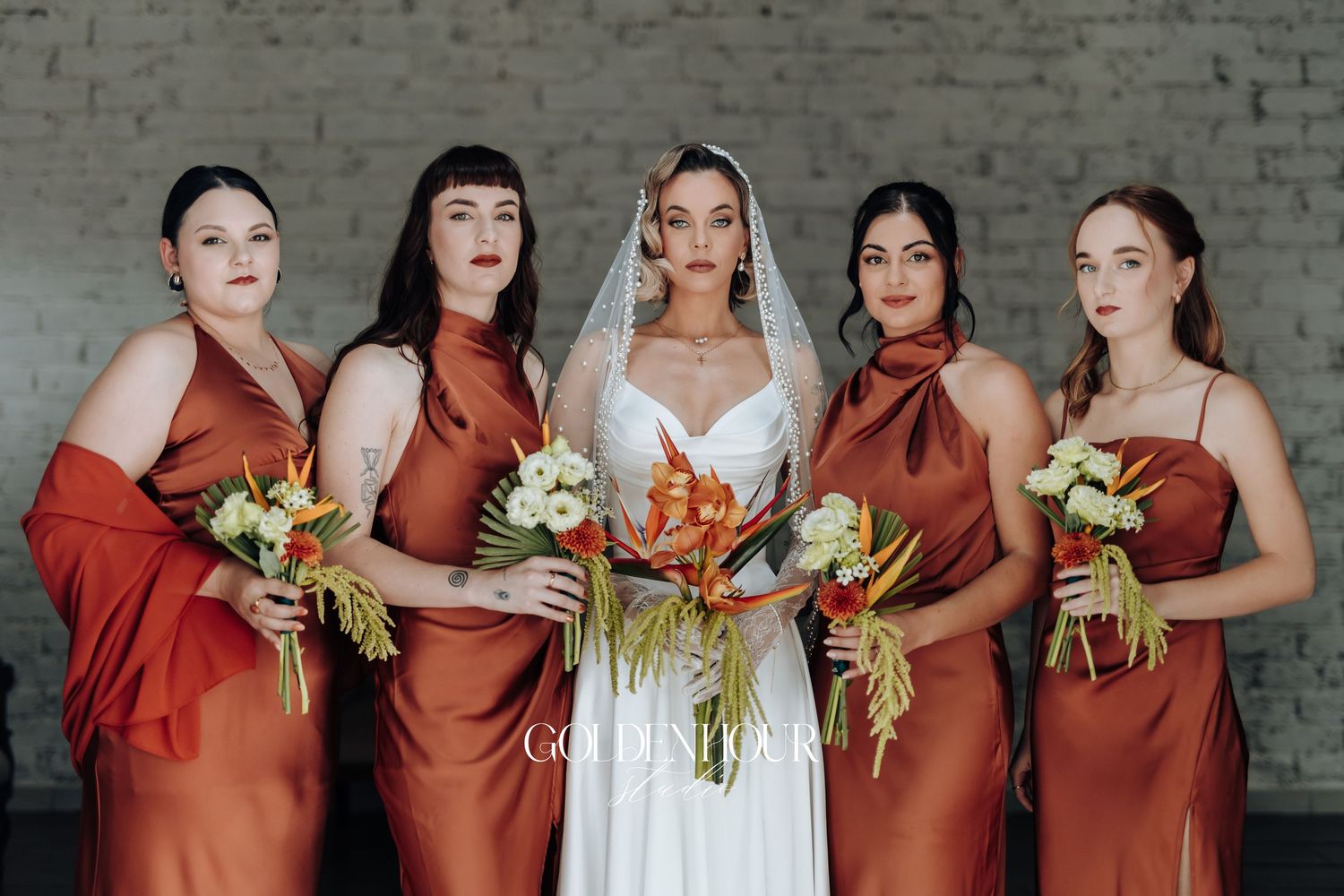 Bridal party in rust-colored dresses posing together against white brick wall.