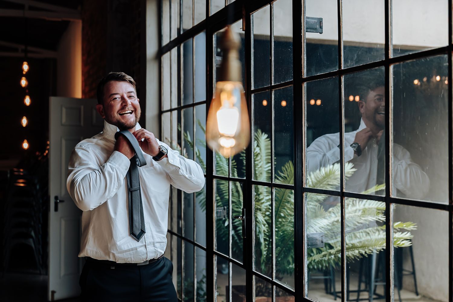 A person in a white dress shirt and black pants stands near a window in a dimly lit room.