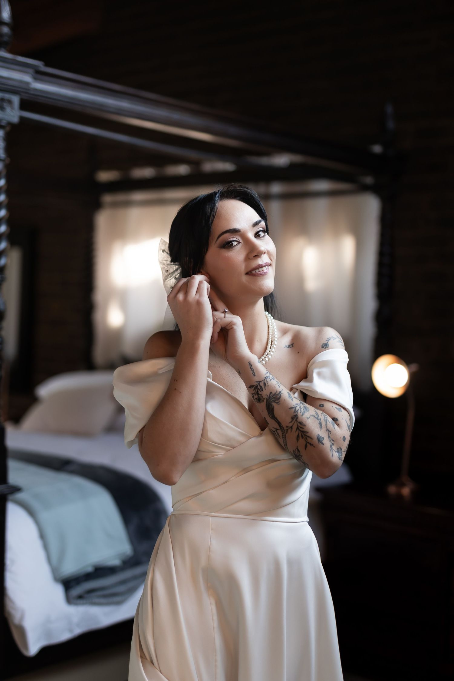 Elegant woman in cream-colored off-shoulder dress with visible arm tattoo puts on jewelry in dimly lit bedroom.