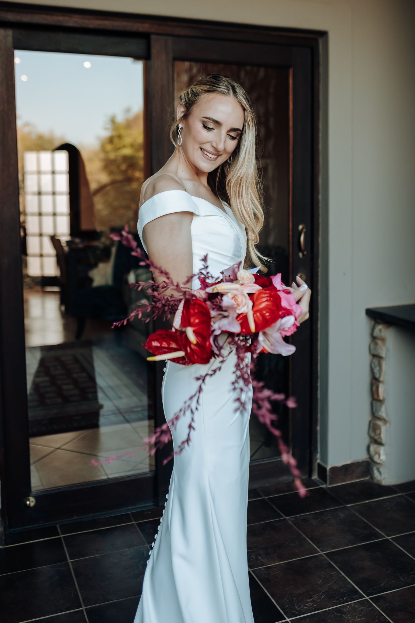 Elegant bride in fitted white dress holding red floral bouquet by glass doors.