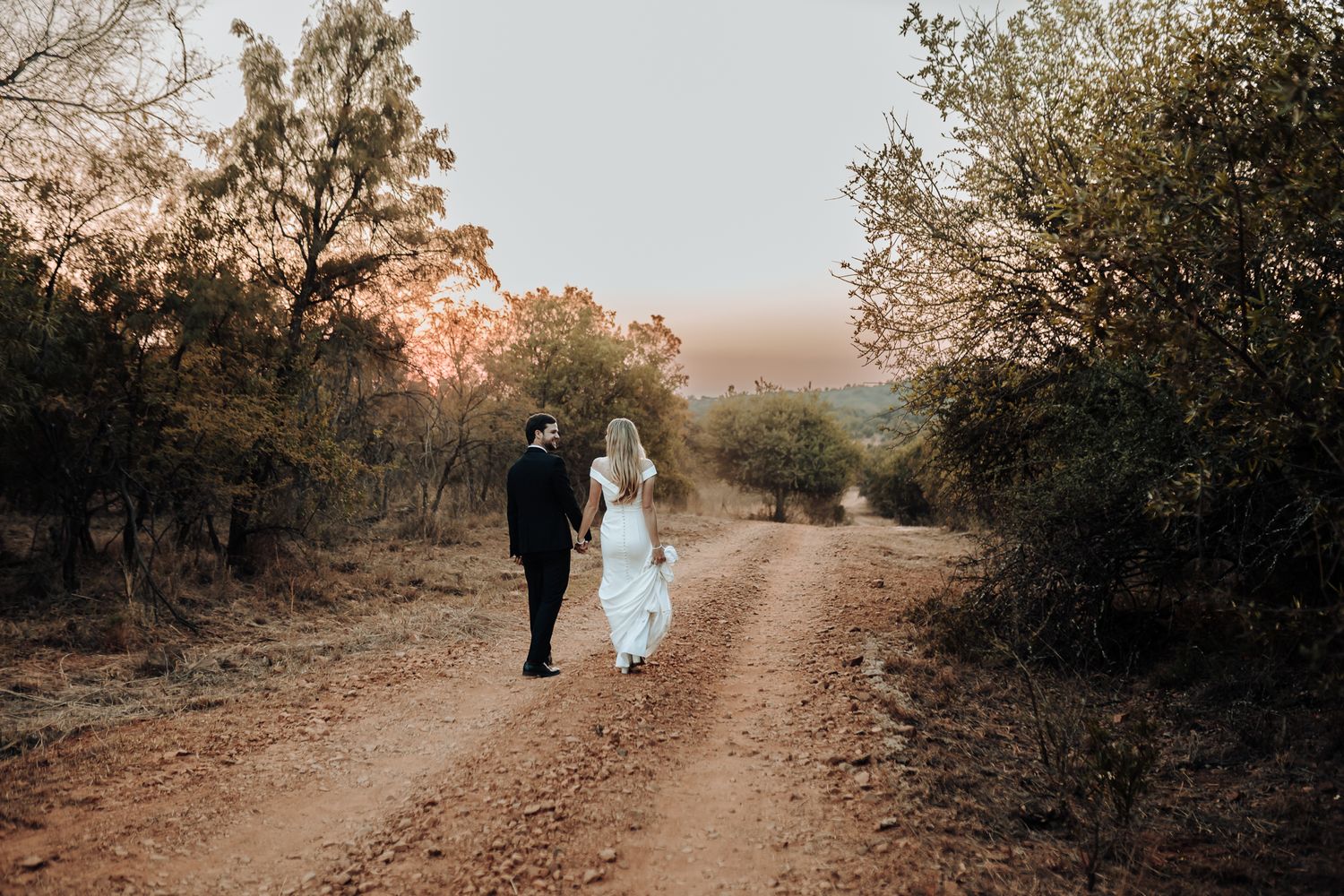 A couple walks hand in hand down a dirt path at sunset surrounded by natural bushland.