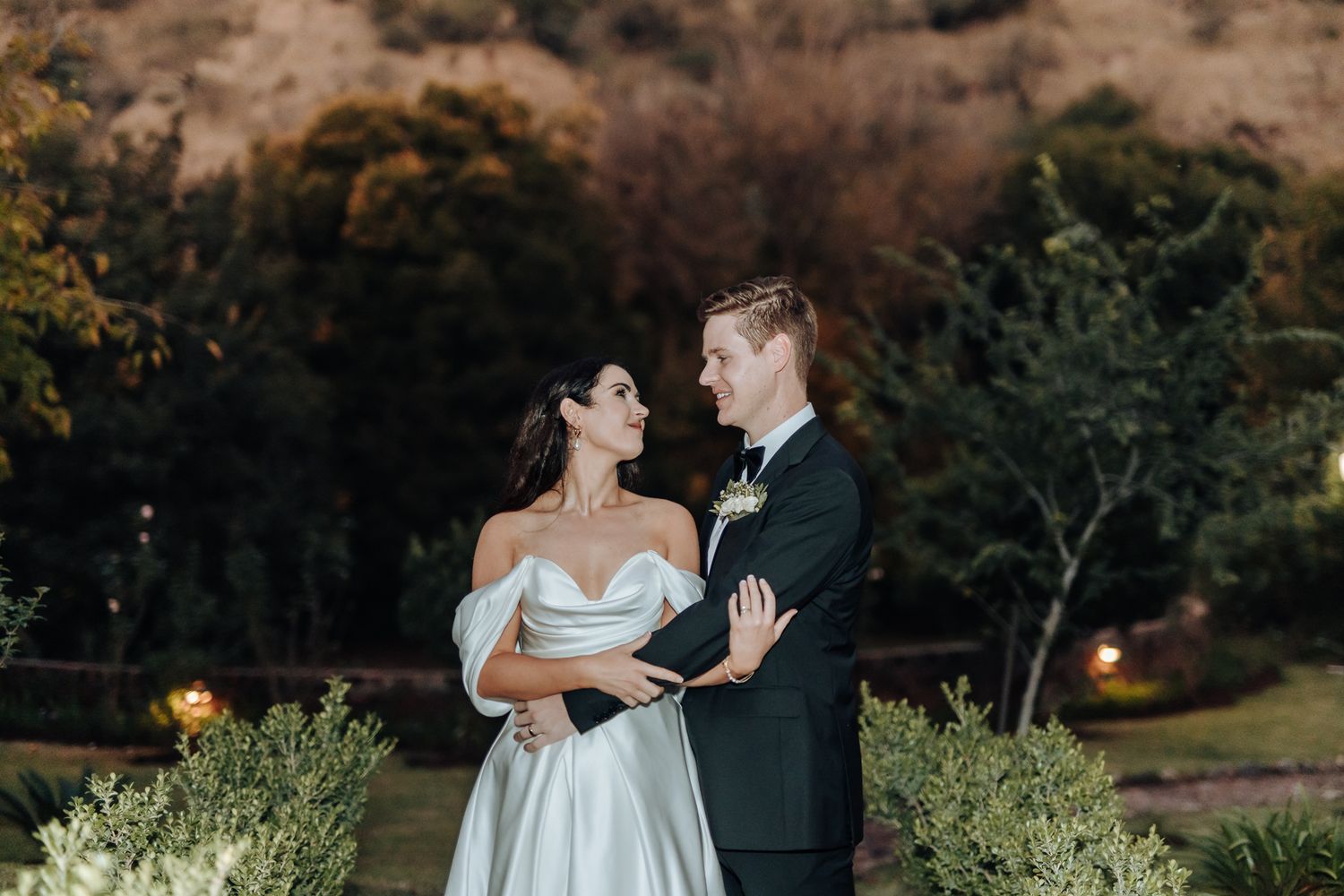 An elegant wedding couple poses in garden with mountains in background at dusk.