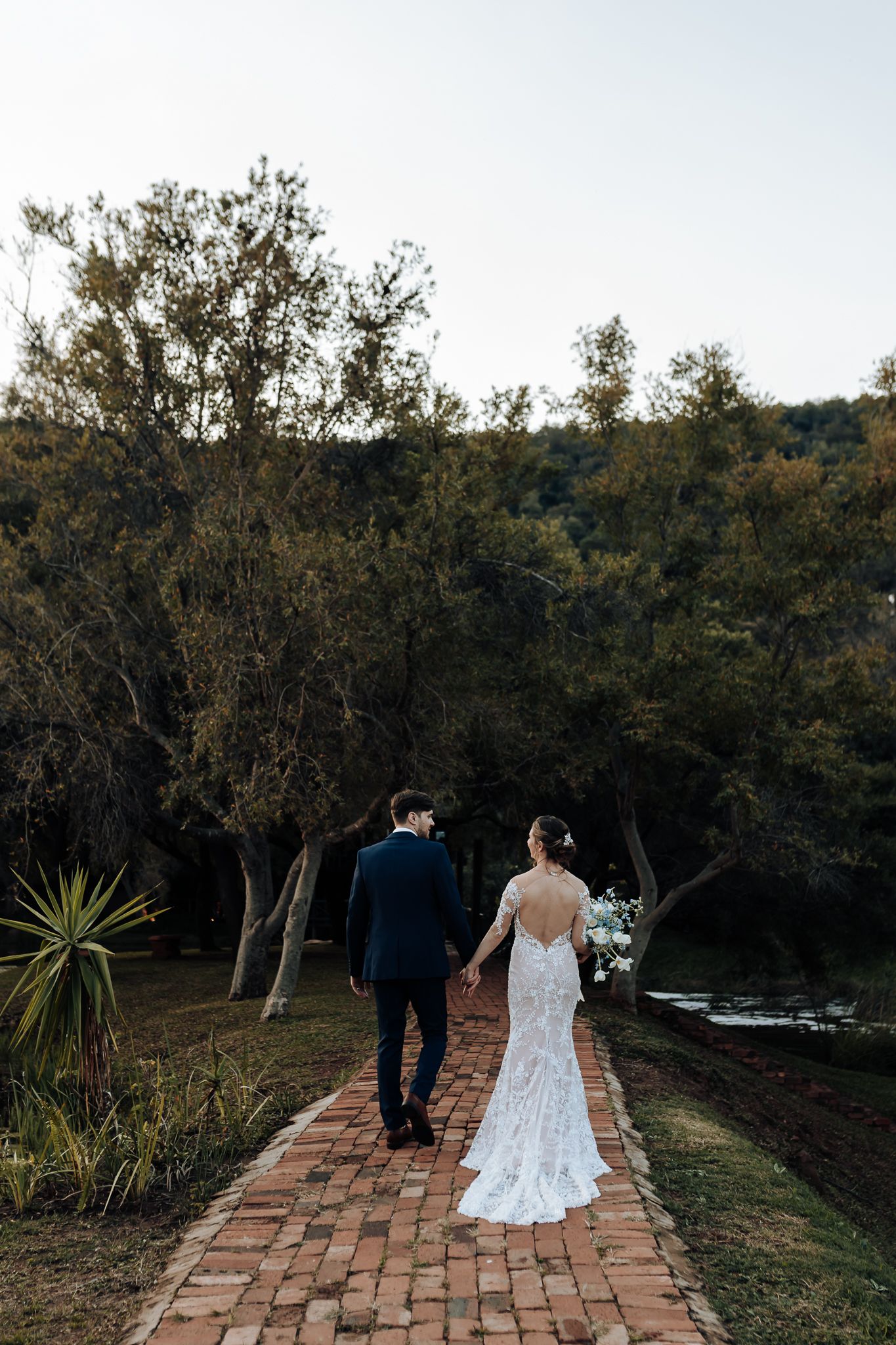 A couple walks along a brick pathway surrounded by lush greenery and water views.