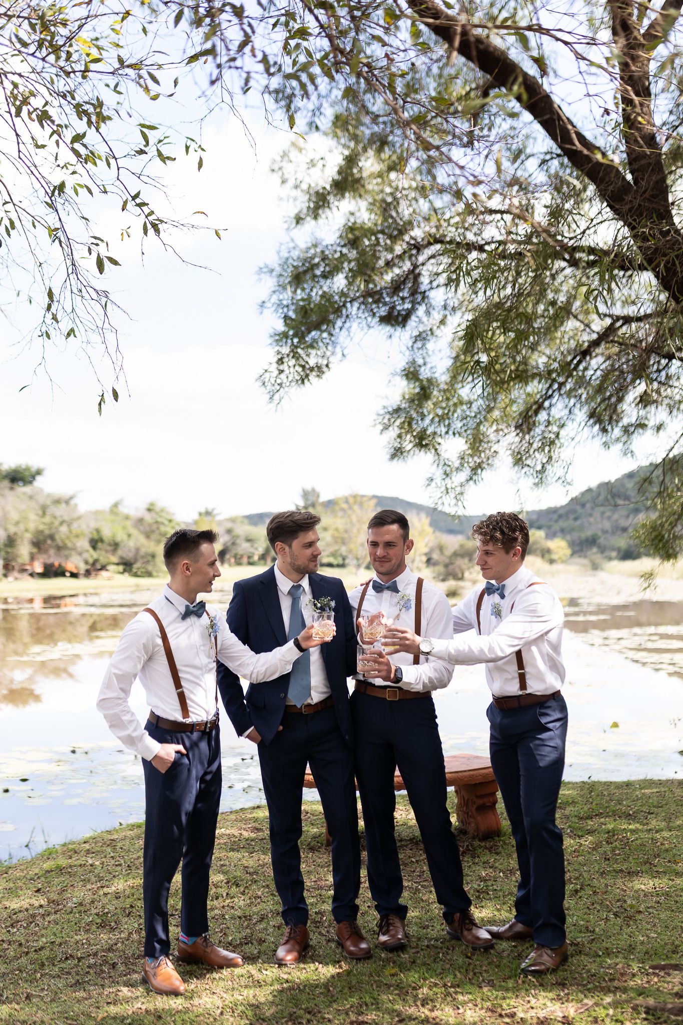 Groomsmen in suspenders and navy pants enjoying drinks by waterfront.