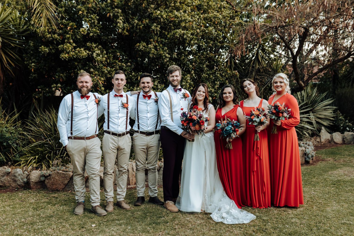 Wedding party posed outdoors with groomsmen in suspenders and bridesmaids in red dresses.