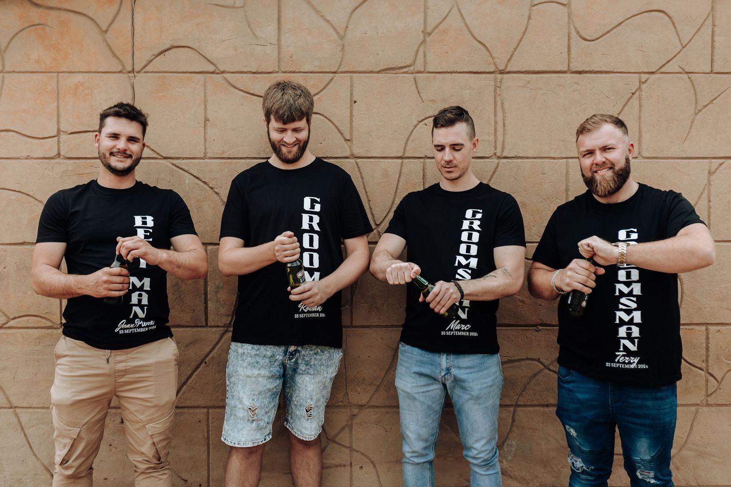 Four friends in matching black t-shirts stand against textured wall holding bottles.
