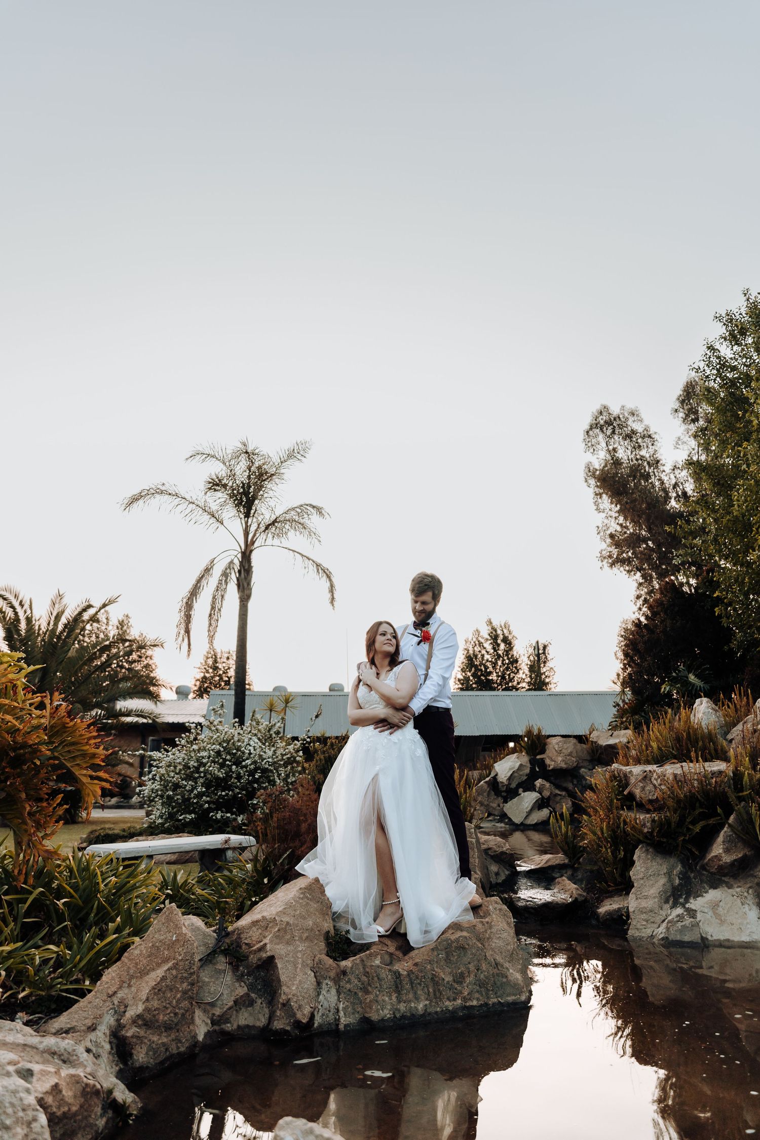 Wedding couple stands atop rock formation near reflecting pool with palm trees at sunset.