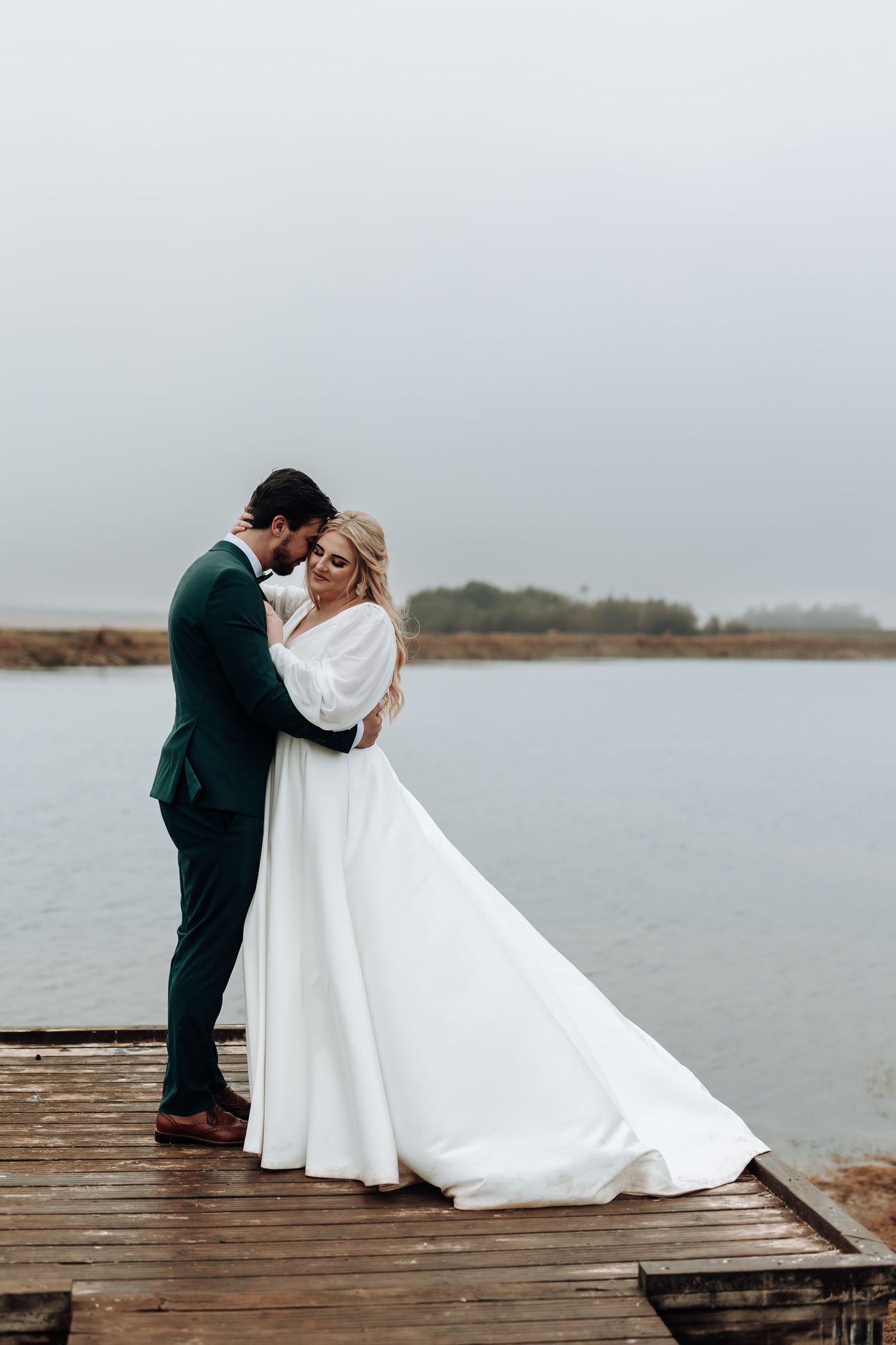 Newlyweds embrace on a lakeside dock with the bride wearing an elegant white gown and groom in emerald suit.