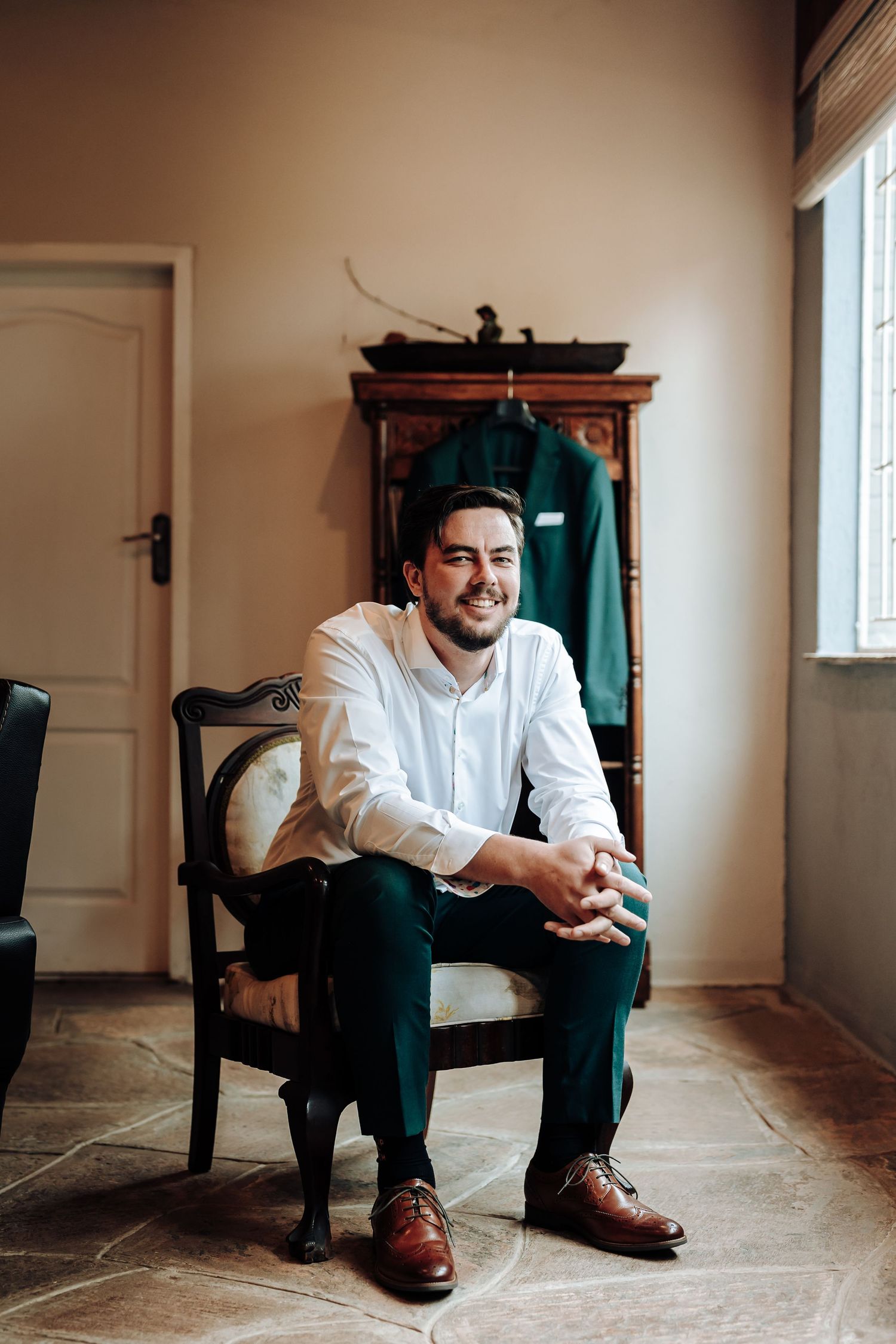 A suited figure sits in an antique chair near a window with a wooden wardrobe in the background.