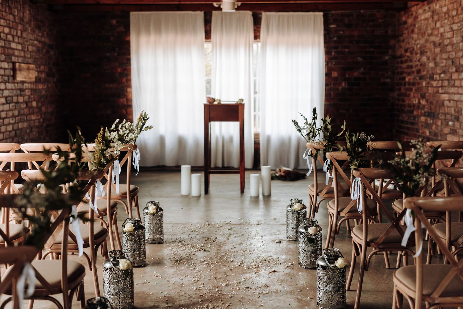 Wedding ceremony setup with wooden chairs and floral decor in a rustic brick room with white curtains.
