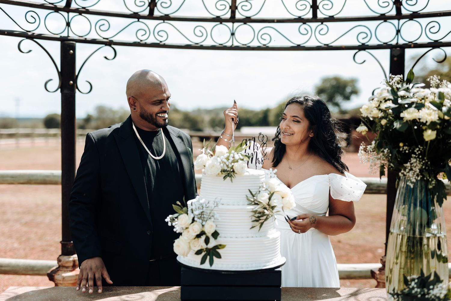 Newlyweds posing under ornate outdoor wedding arch with white floral decorations.