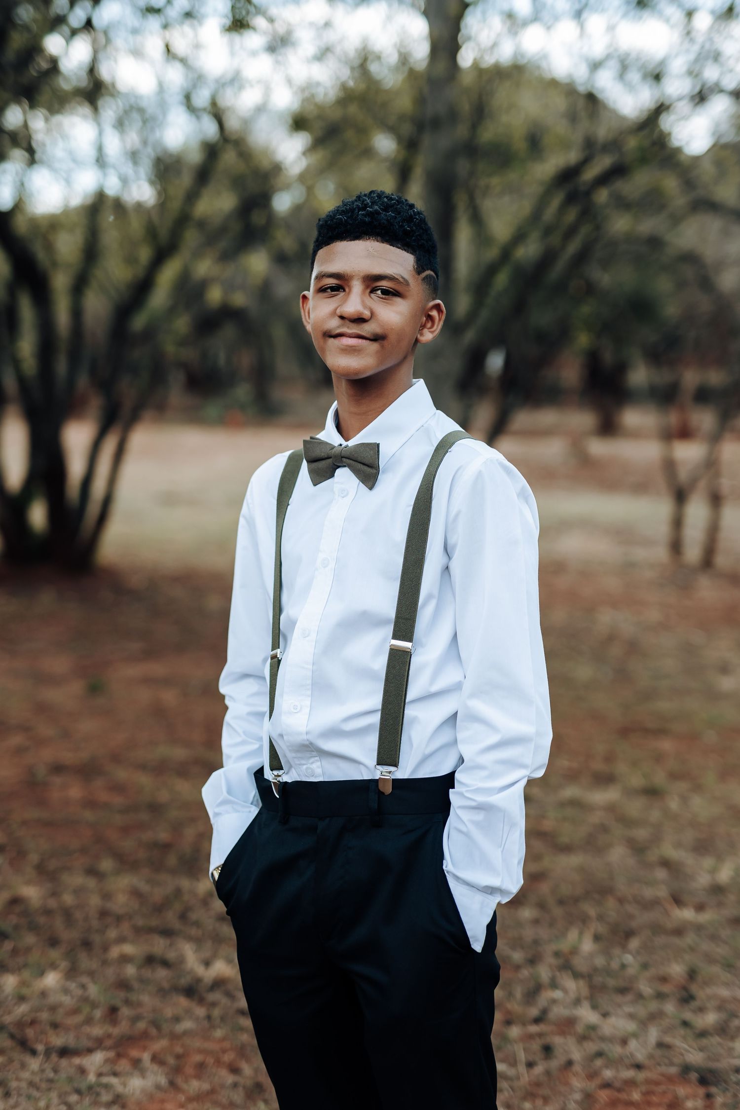 Young man in white dress shirt, bow tie and suspenders smiles in outdoor setting.