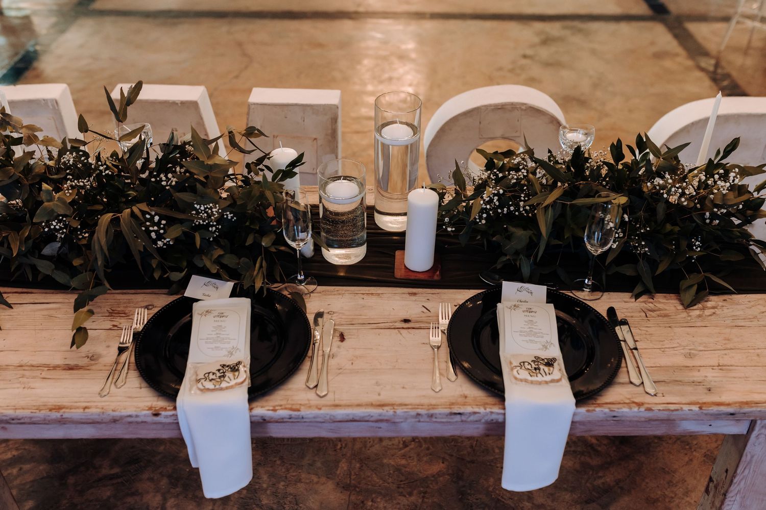 Rustic wedding reception table with black plates and lush greenery centerpieces.