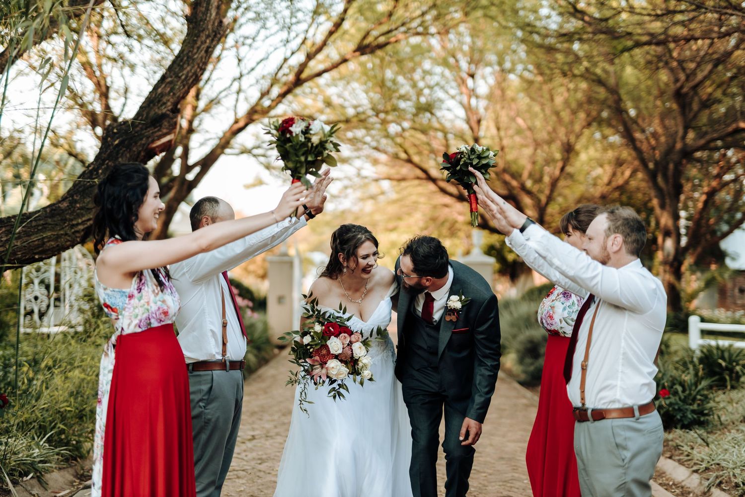 Wedding party members raise bouquets in celebration on a tree-lined pathway.