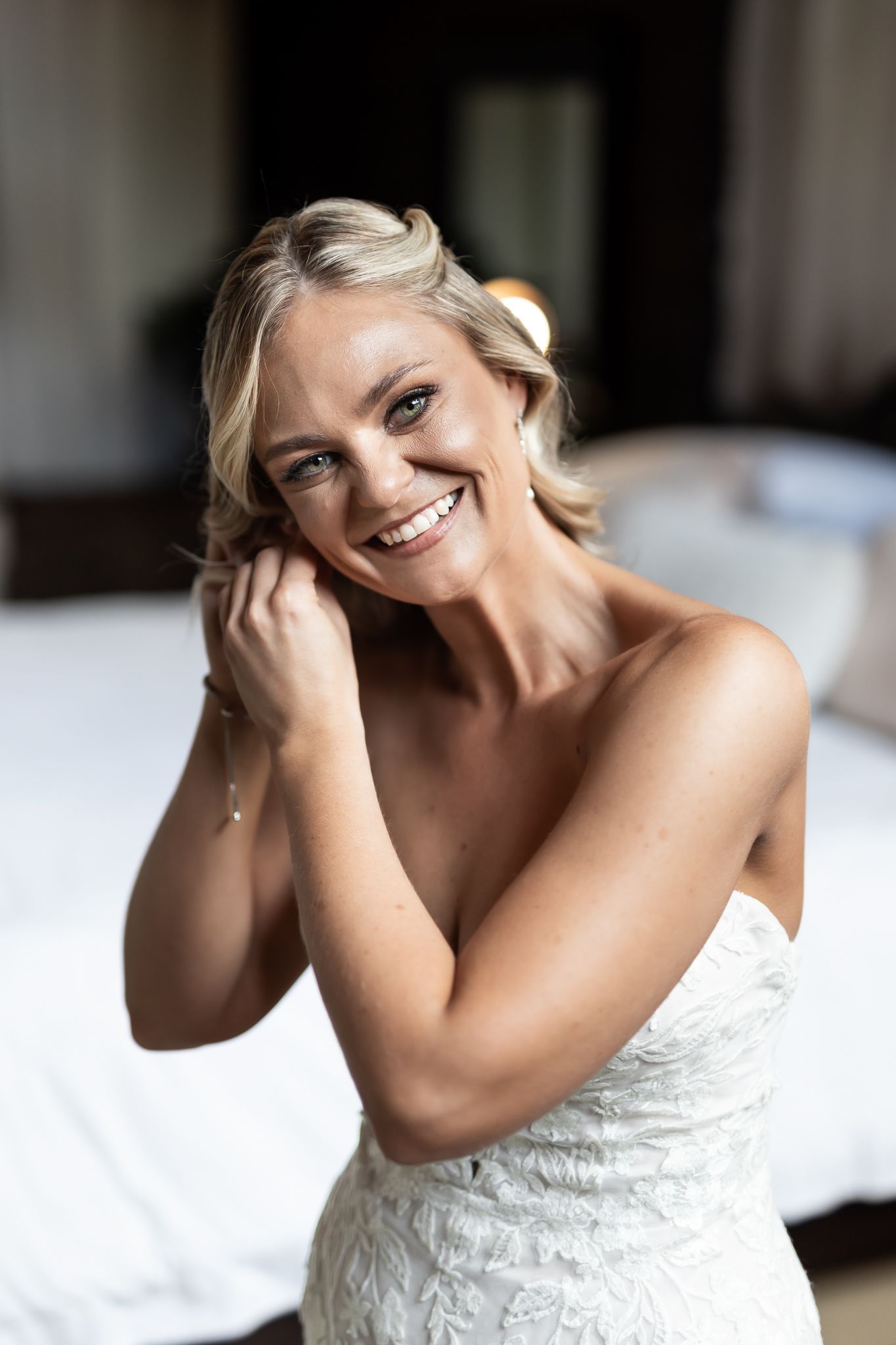 Radiant bride in strapless wedding dress smiles while posing in bedroom setting.