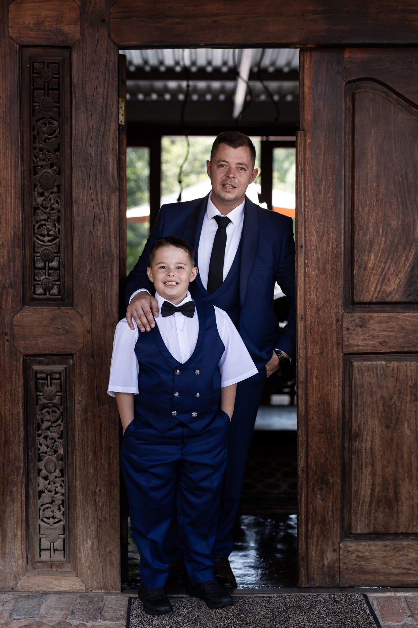 A formal portrait of two people in suits standing in an ornate wooden doorway.