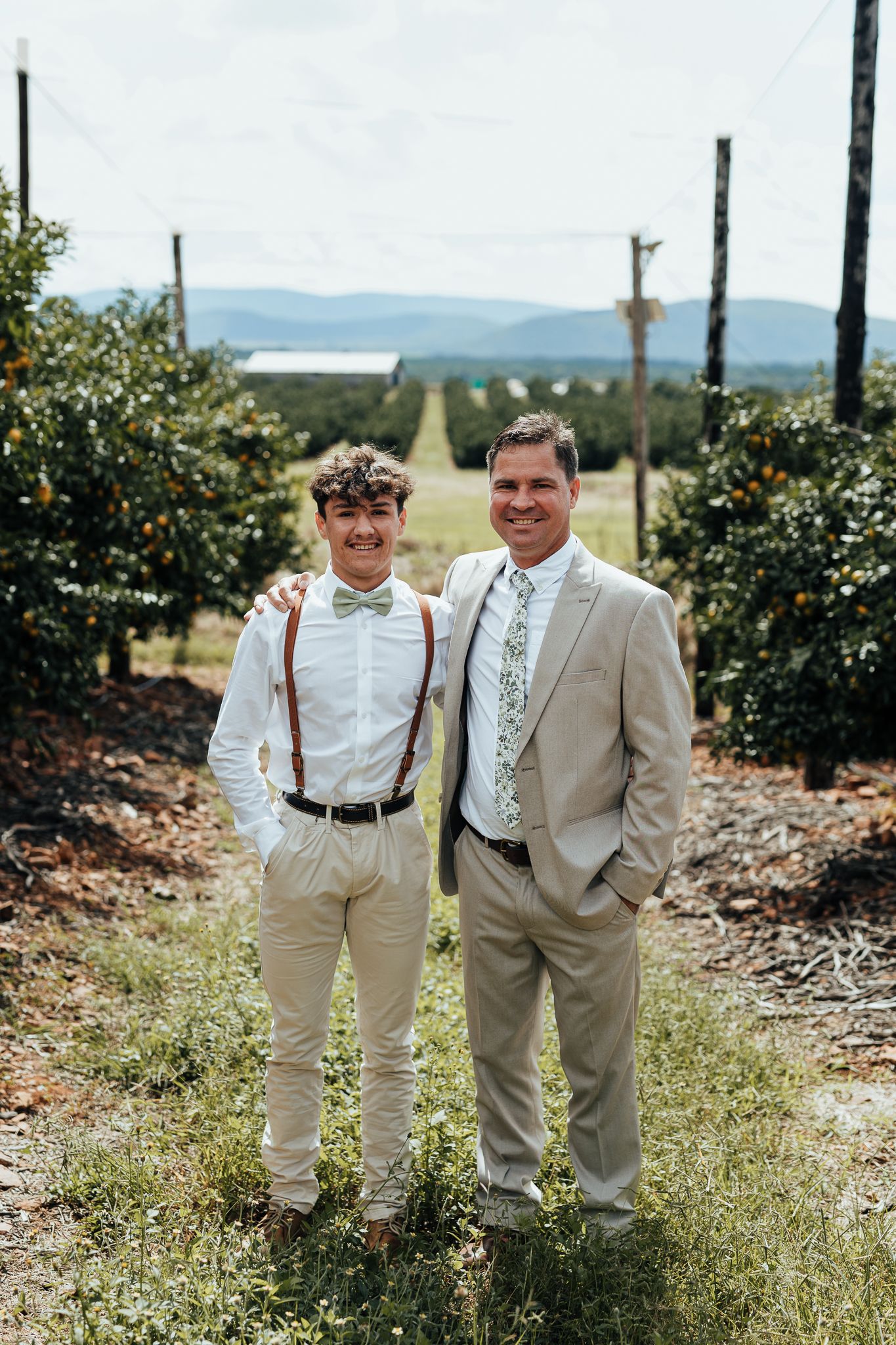 Two wedding attendees in light colored suits pose together in an orchard with mountains in the background.