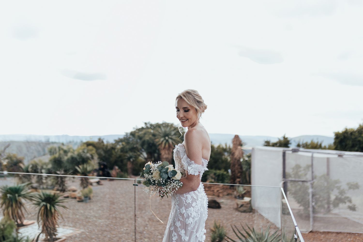 Bride in detailed lace gown poses with bouquet on modern outdoor terrace.