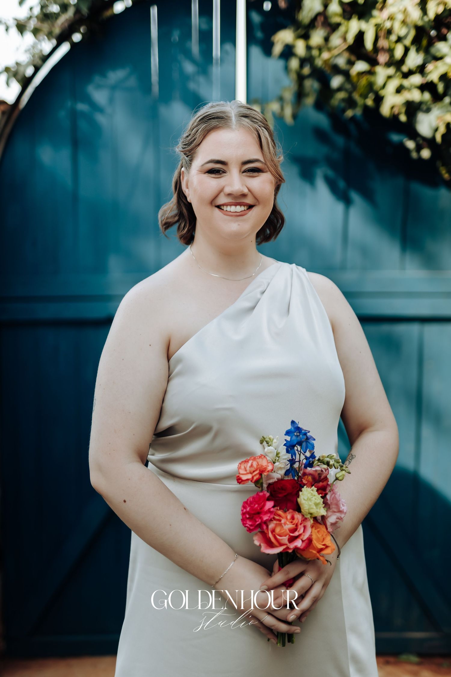 A person in an elegant one-shoulder cream dress holds a vibrant bouquet with pink and blue flowers against a teal backdrop.