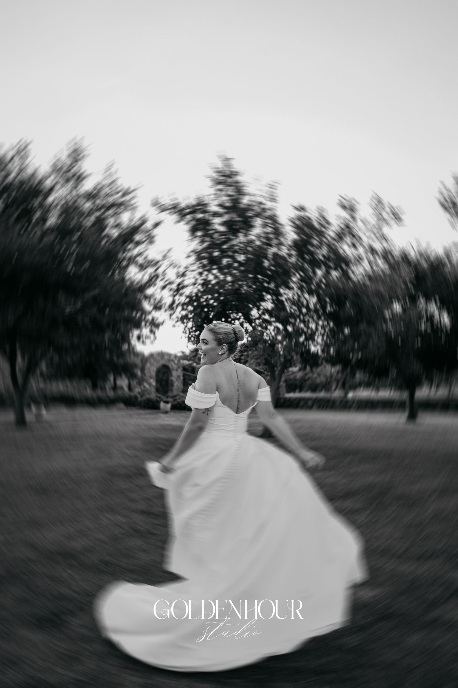 Black and white motion-blurred photo of someone in a flowing white wedding dress spinning in an outdoor garden setting.