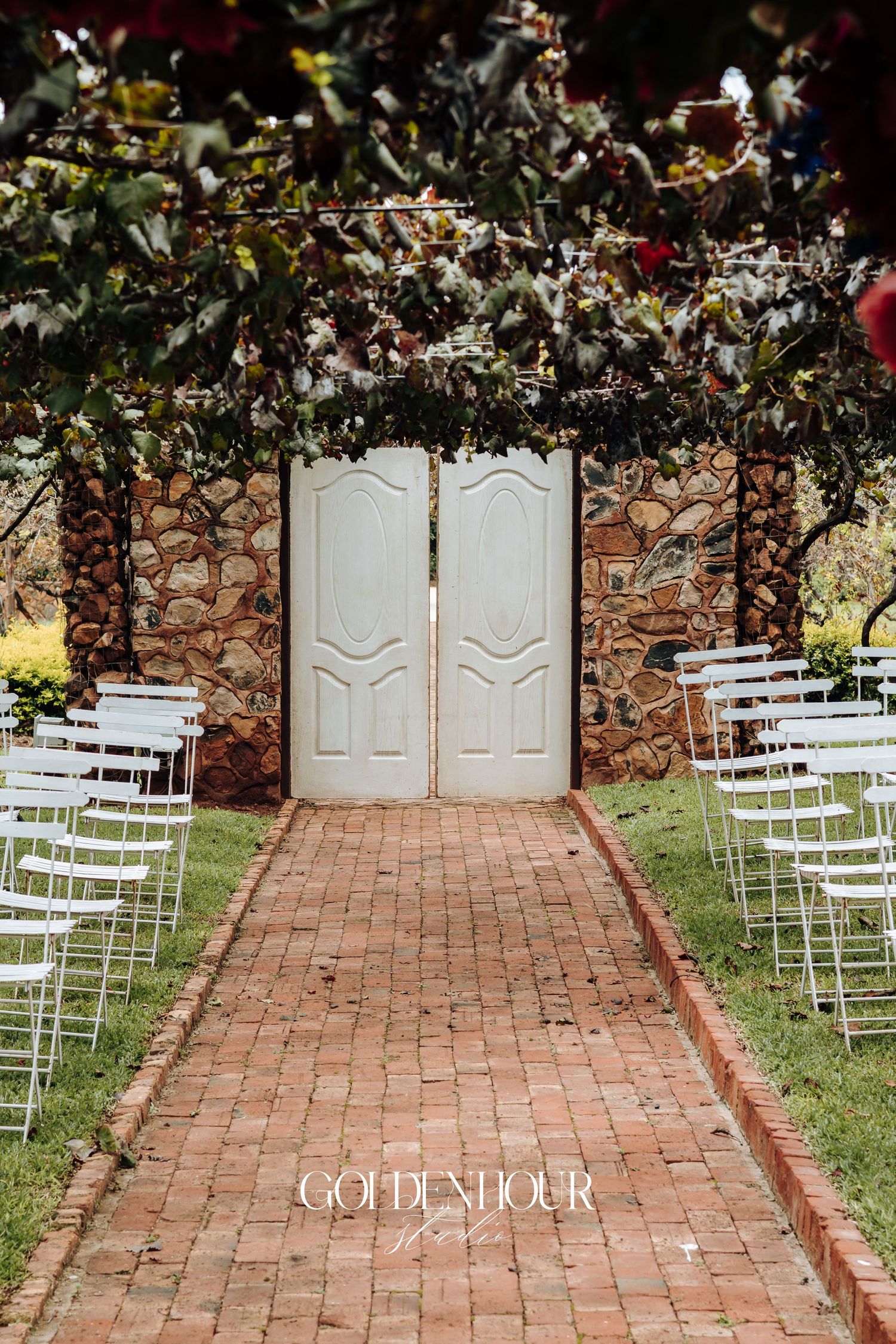 White garden chairs arranged for an outdoor wedding ceremony on brick pavers with vintage white doors and floral arch.