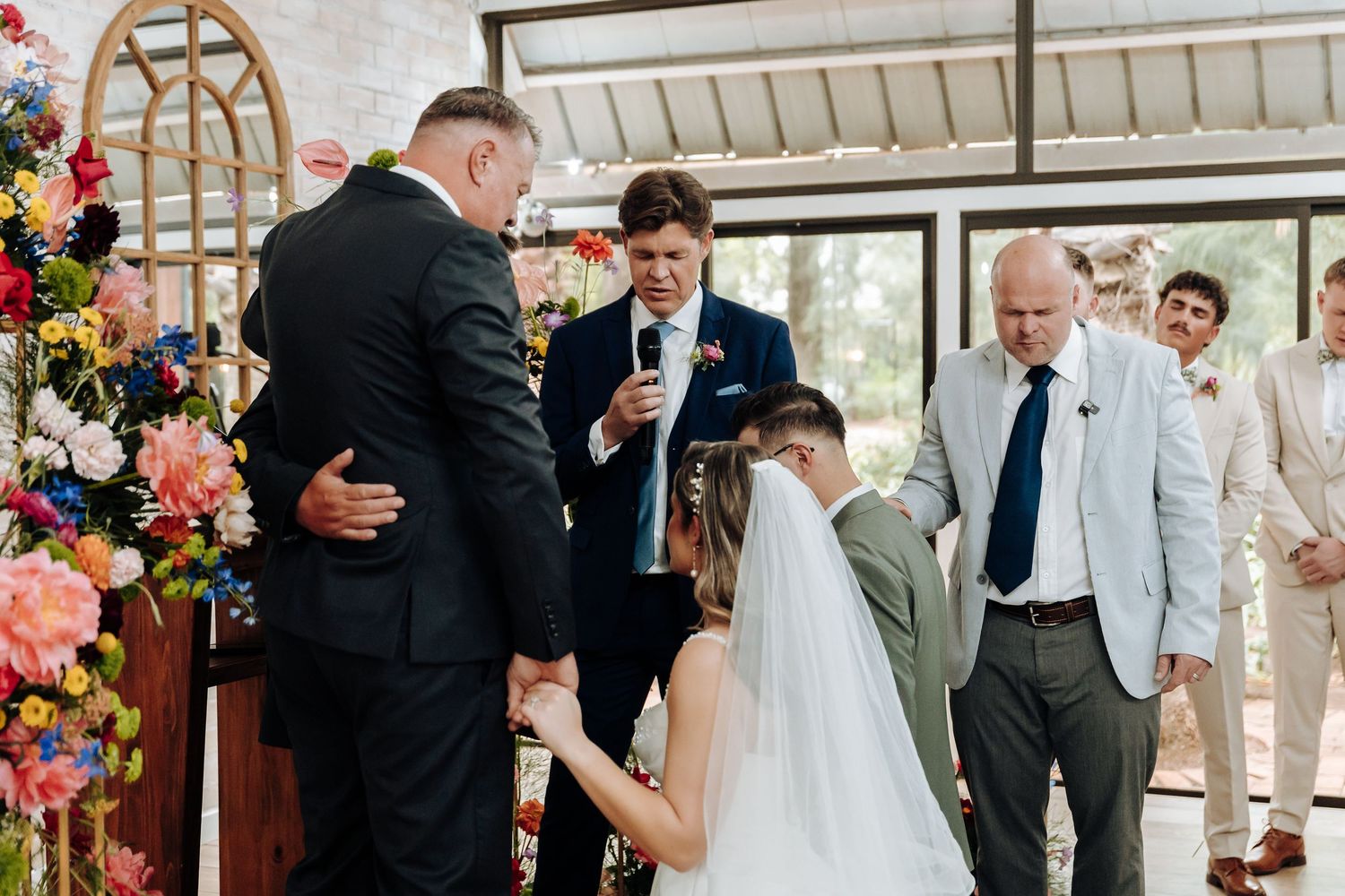 Wedding ceremony in a bright indoor venue with floral arrangements and guests watching.