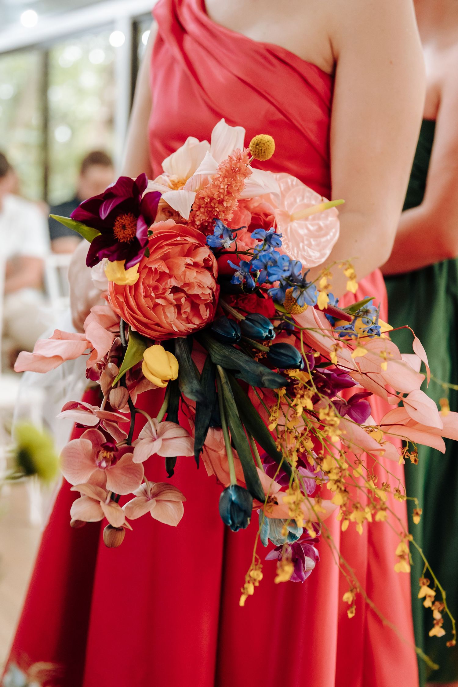 Vibrant wedding bouquet with coral peonies, blue delphiniums, and orchids against a red bridesmaid dress.