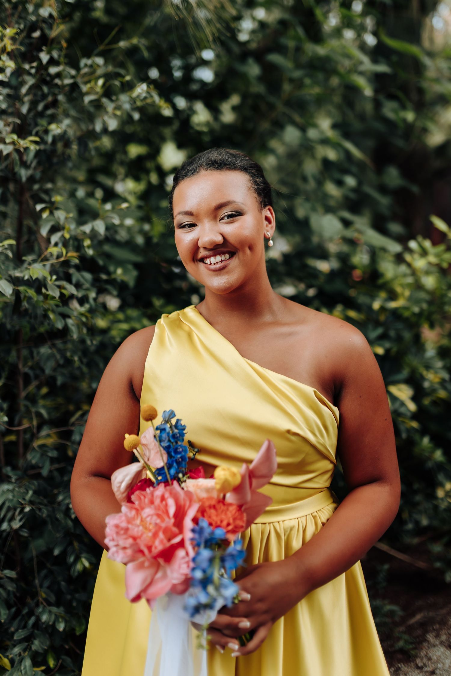 A person wearing a yellow one-shoulder dress holds vibrant blue and coral flowers in an outdoor setting.