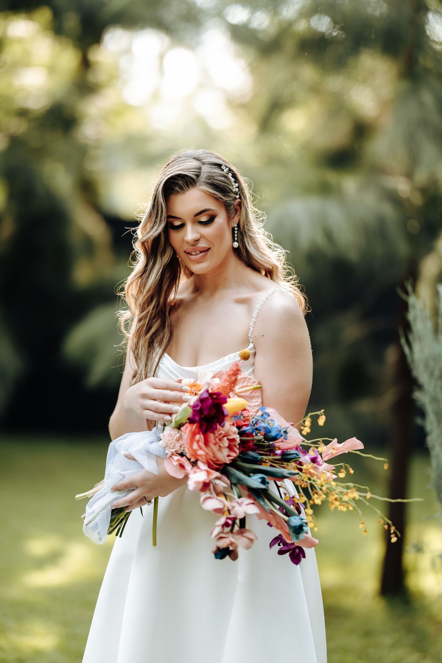 Bride in white off-shoulder dress holding colorful autumn floral bouquet in sunlit garden setting.