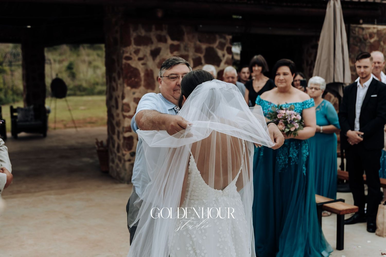 Wedding party in teal blue dresses and formal attire gather outdoors for ceremony under natural lighting.