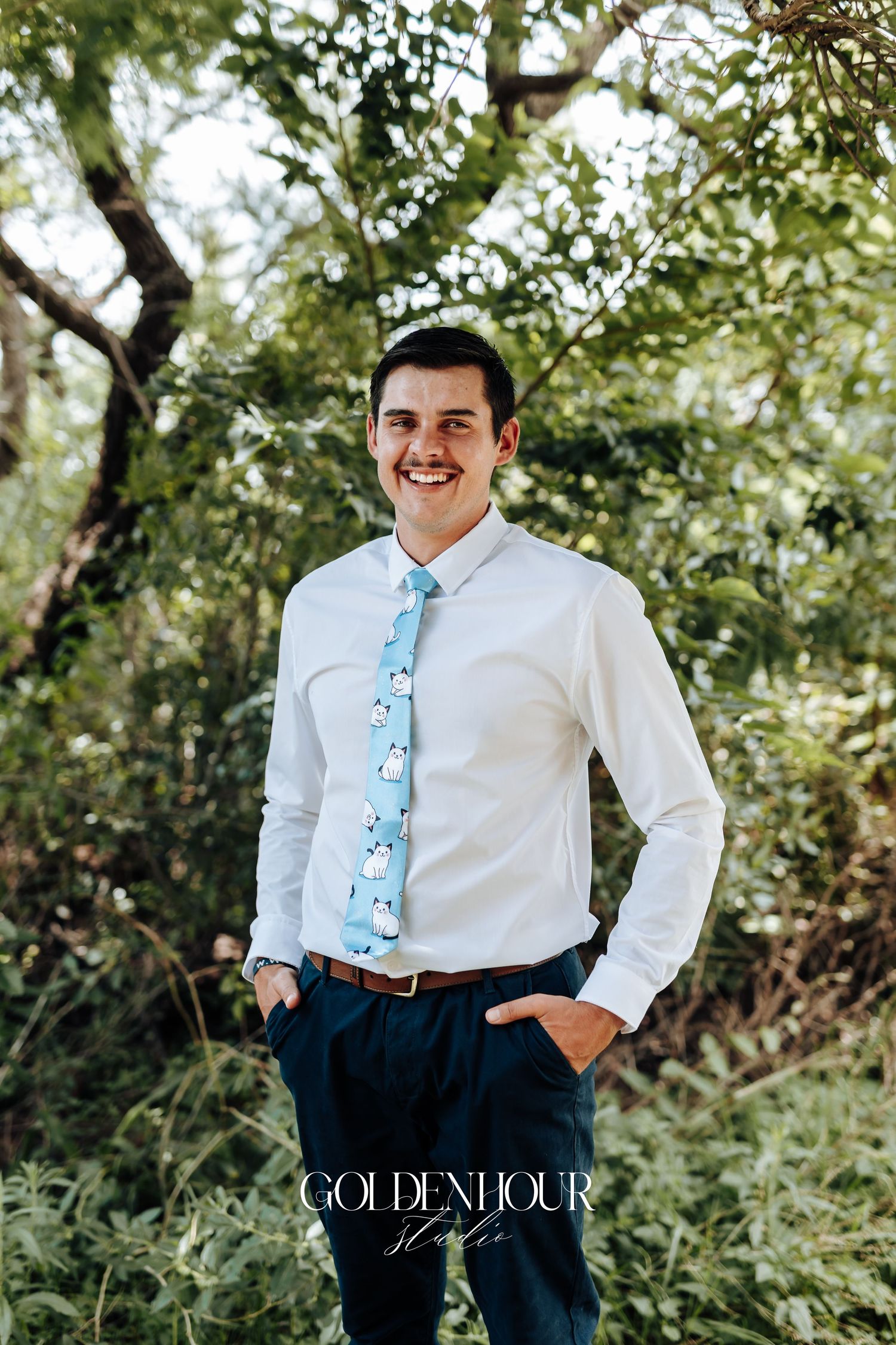 A groom in a white dress shirt and floral blue tie poses outdoors against a leafy green background.