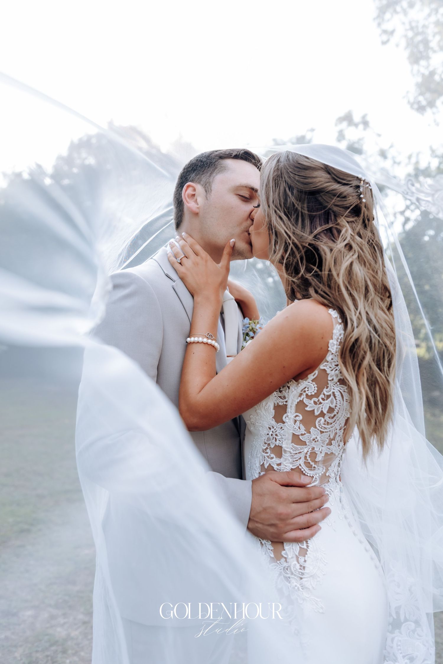 Romantic wedding couple embraces in an ethereal outdoor setting with flowing white veil and lace dress details.