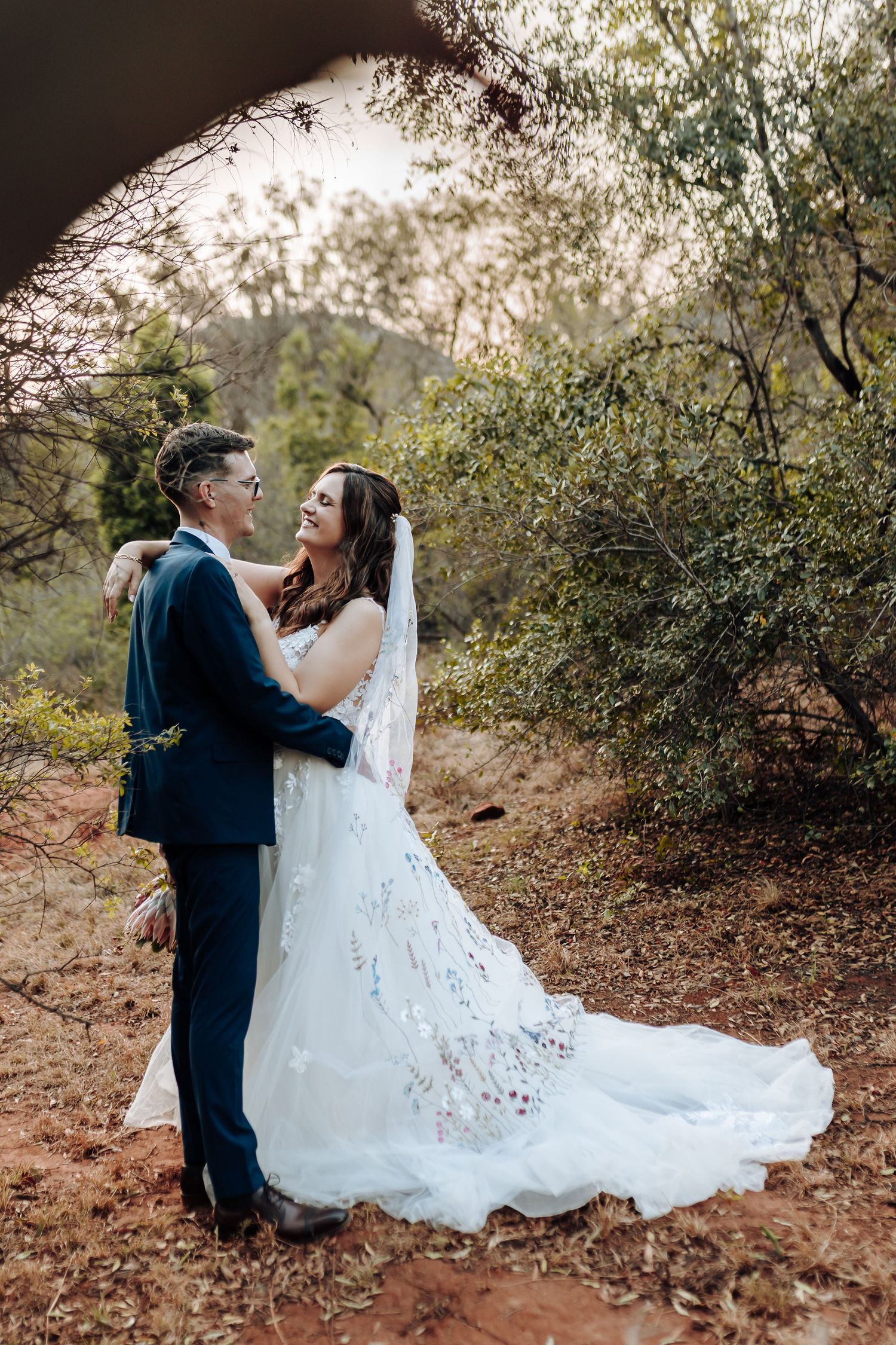 A couple in wedding attire share a romantic moment together on a wooded path during golden hour.