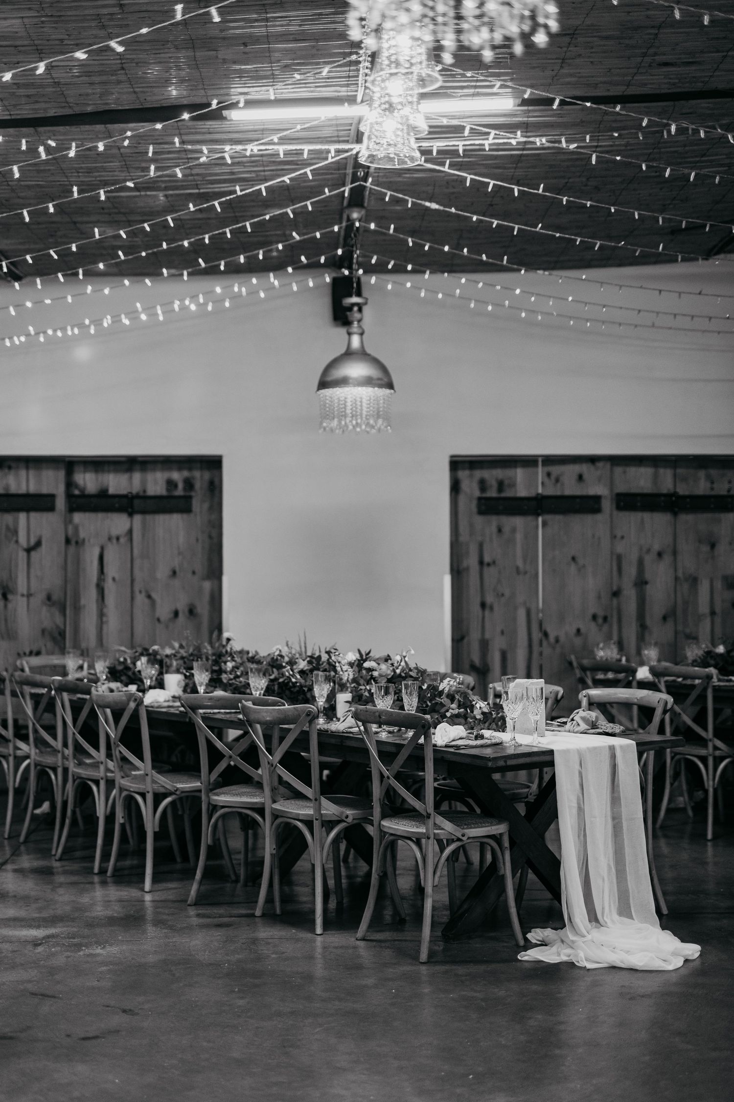 Black and white photo of an elegant wedding reception venue with string lights and rustic wooden tables arranged in rows.
