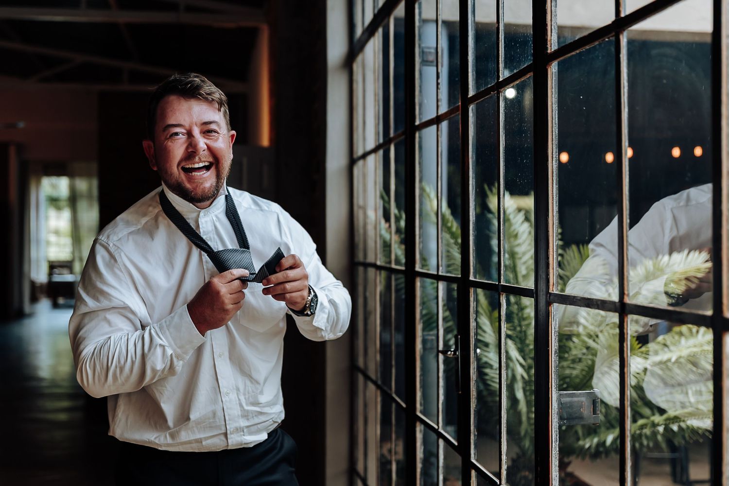 A person in a white dress shirt laughs joyfully while standing near a window with string lights in the background.