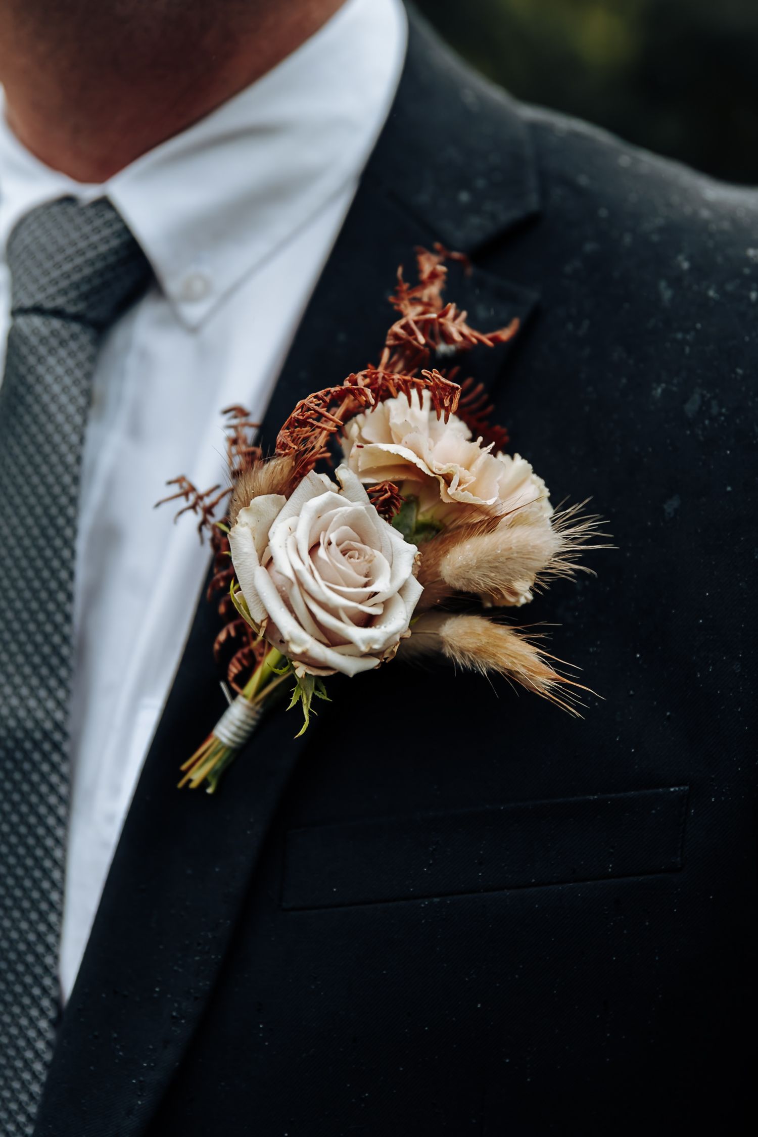 Close-up details of a beige rose and dried flower boutonniere pinned to a black suit jacket with gray tie.
