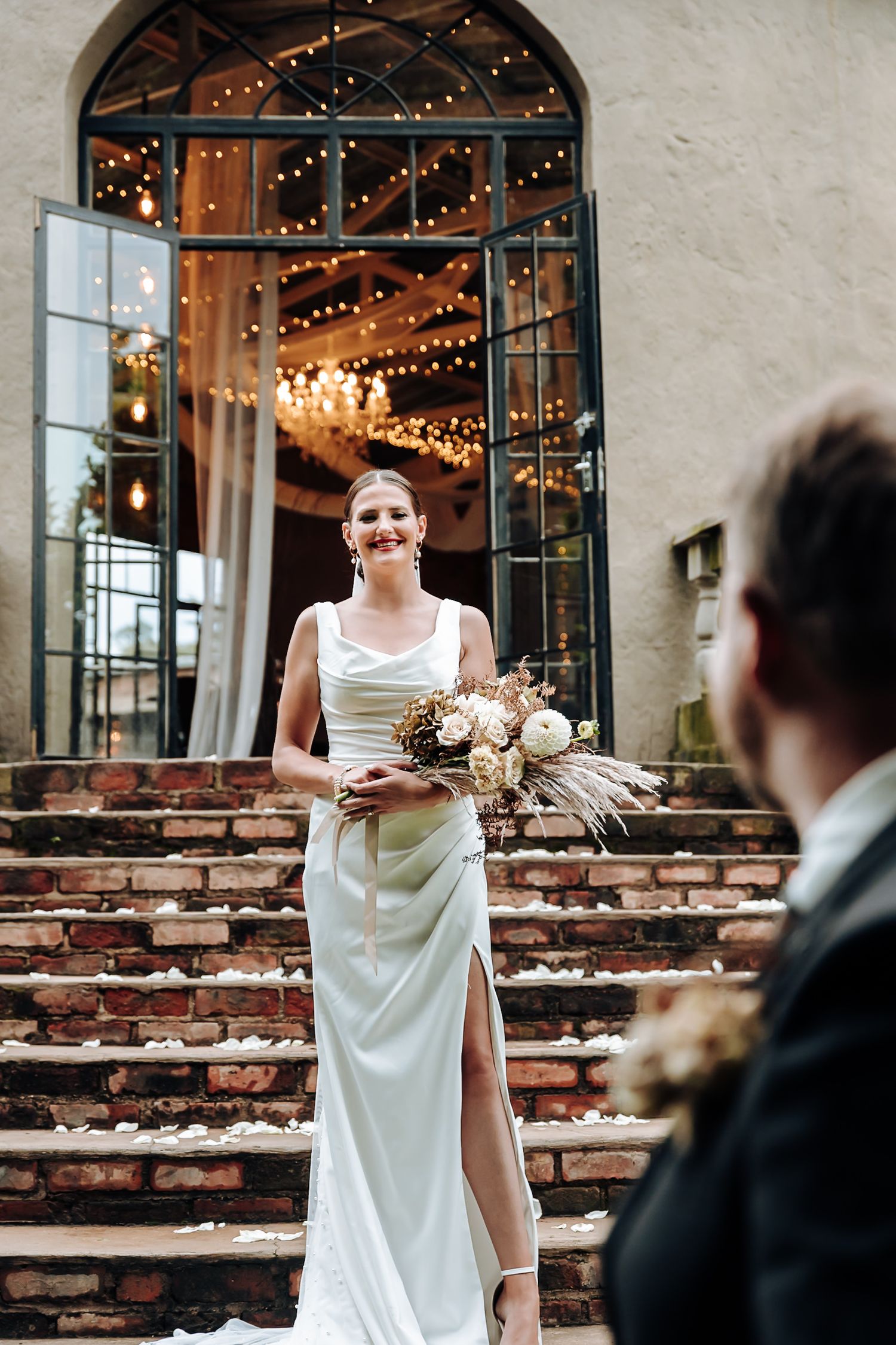 A bride in a flowing white dress stands on rustic brick steps outside a venue with arched windows and string lights.