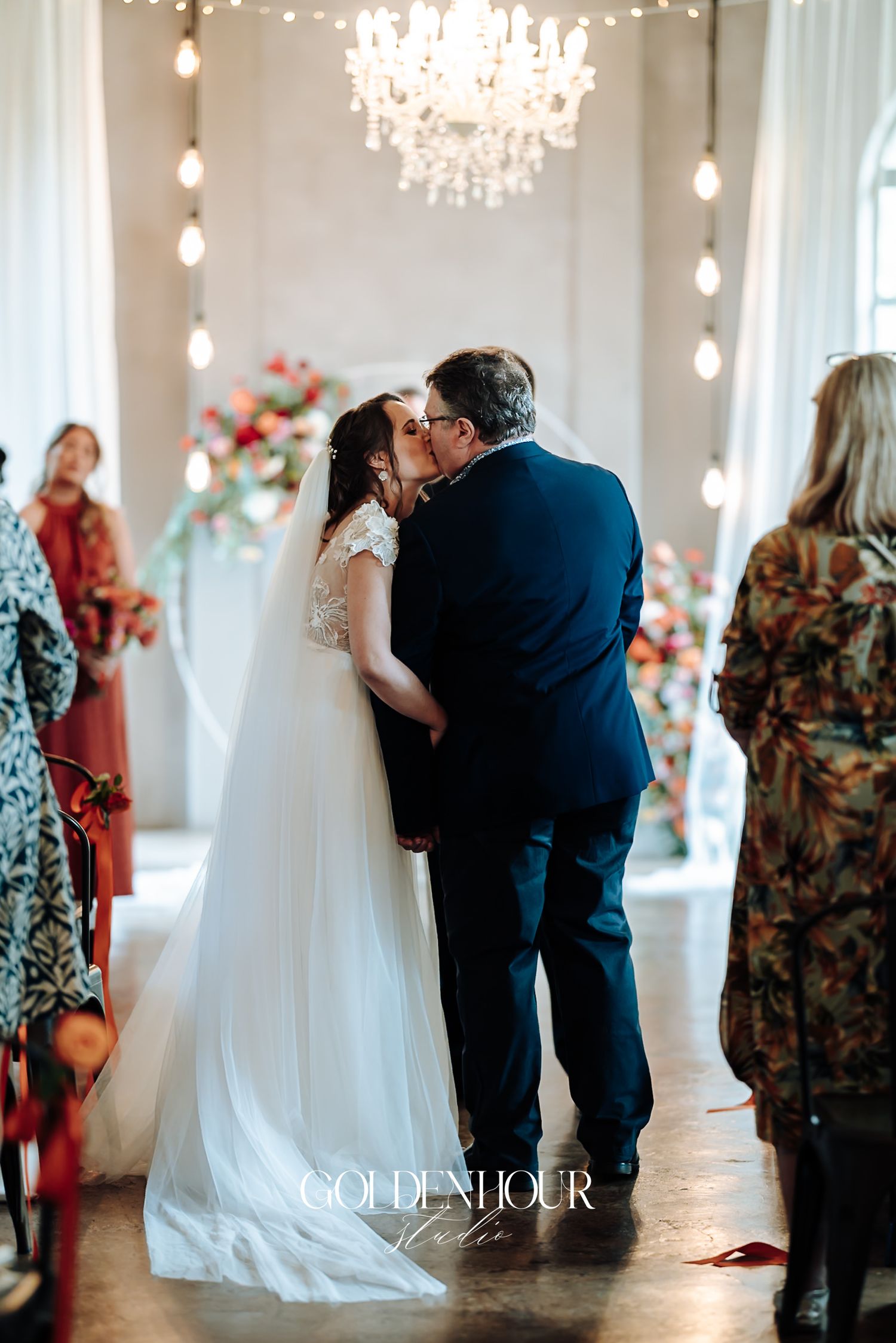 Bride and groom sharing first kiss at altar under chandelier in elegant wedding ceremony venue.