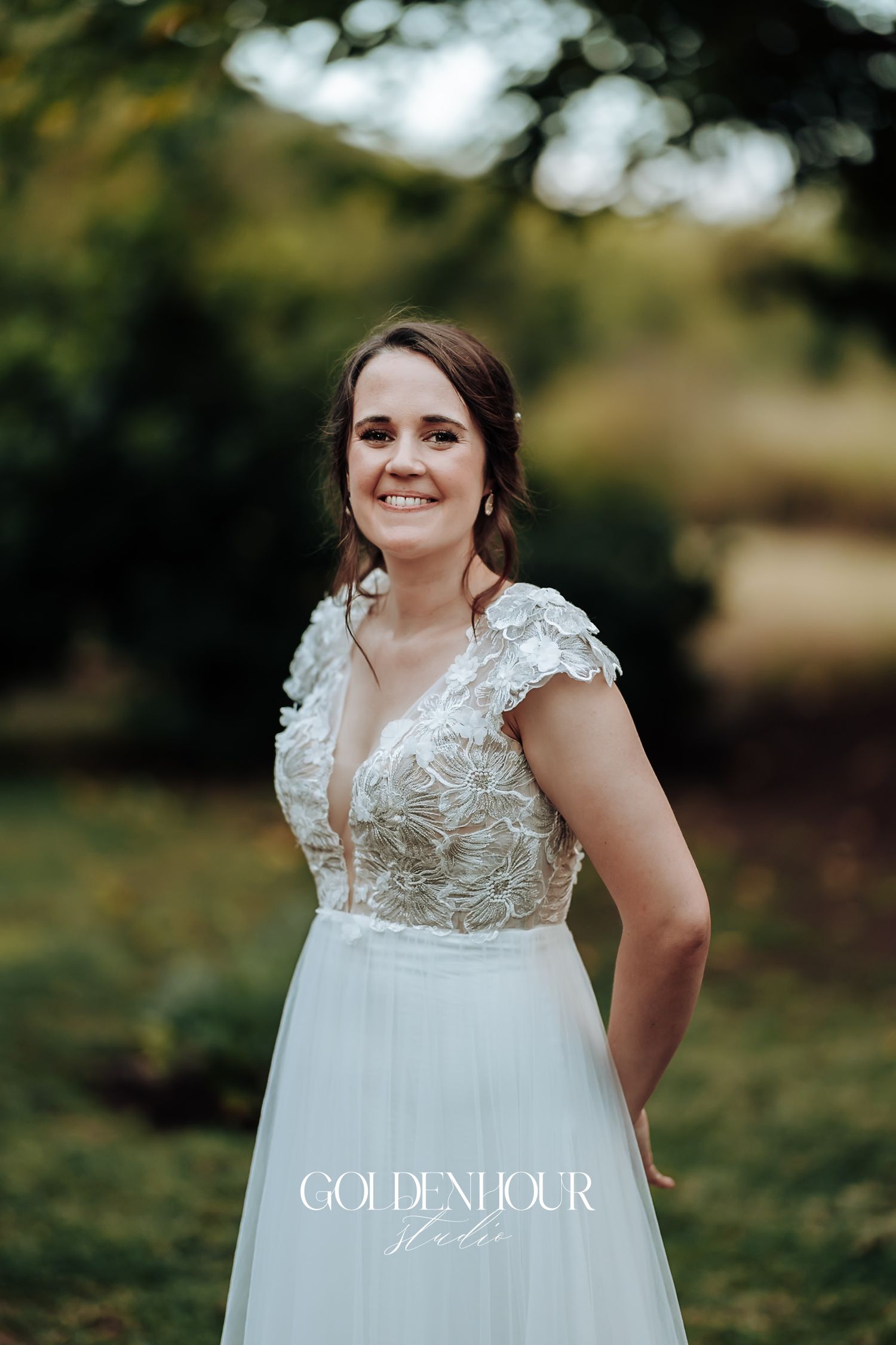 Radiant bride in lace cap sleeve wedding gown smiling in soft golden outdoor garden light.