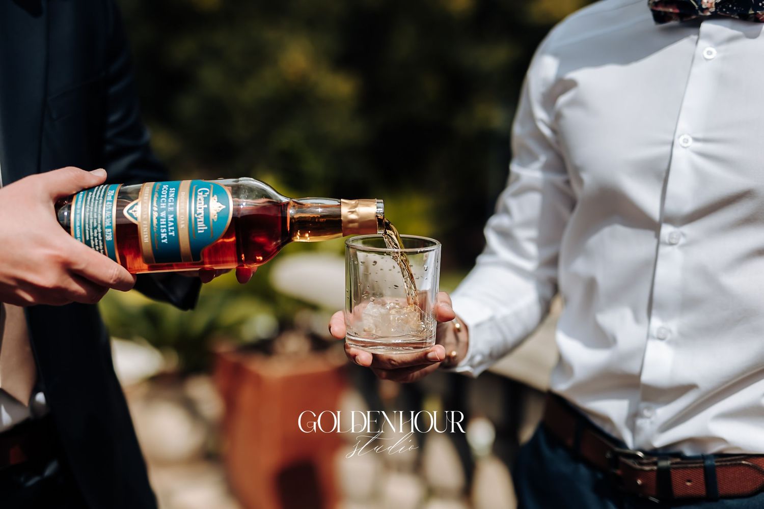Whiskey being poured into a glass held by a groomsman during wedding celebrations outdoors.