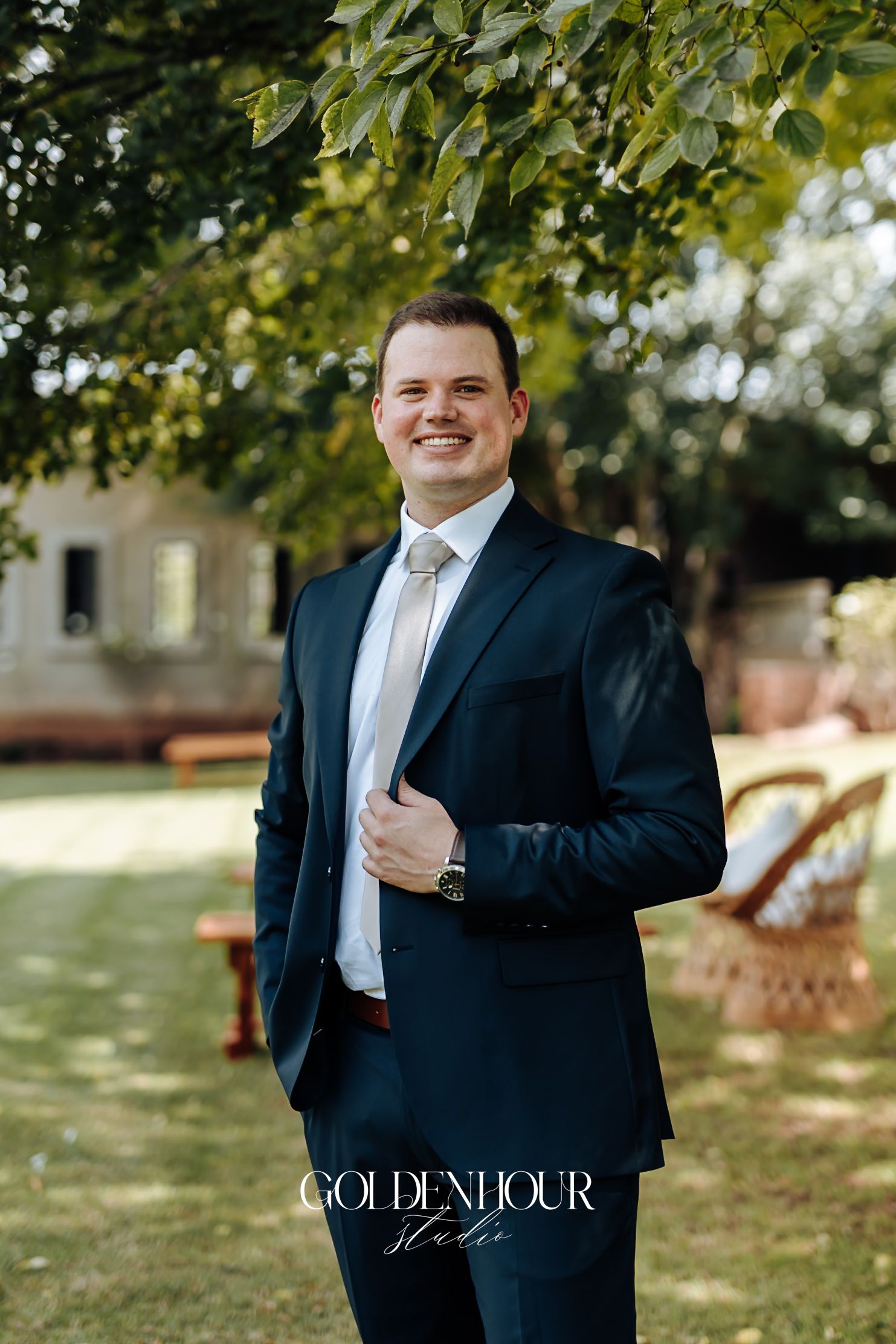Groom in navy suit and white tie smiles confidently outdoors at his wedding venue.