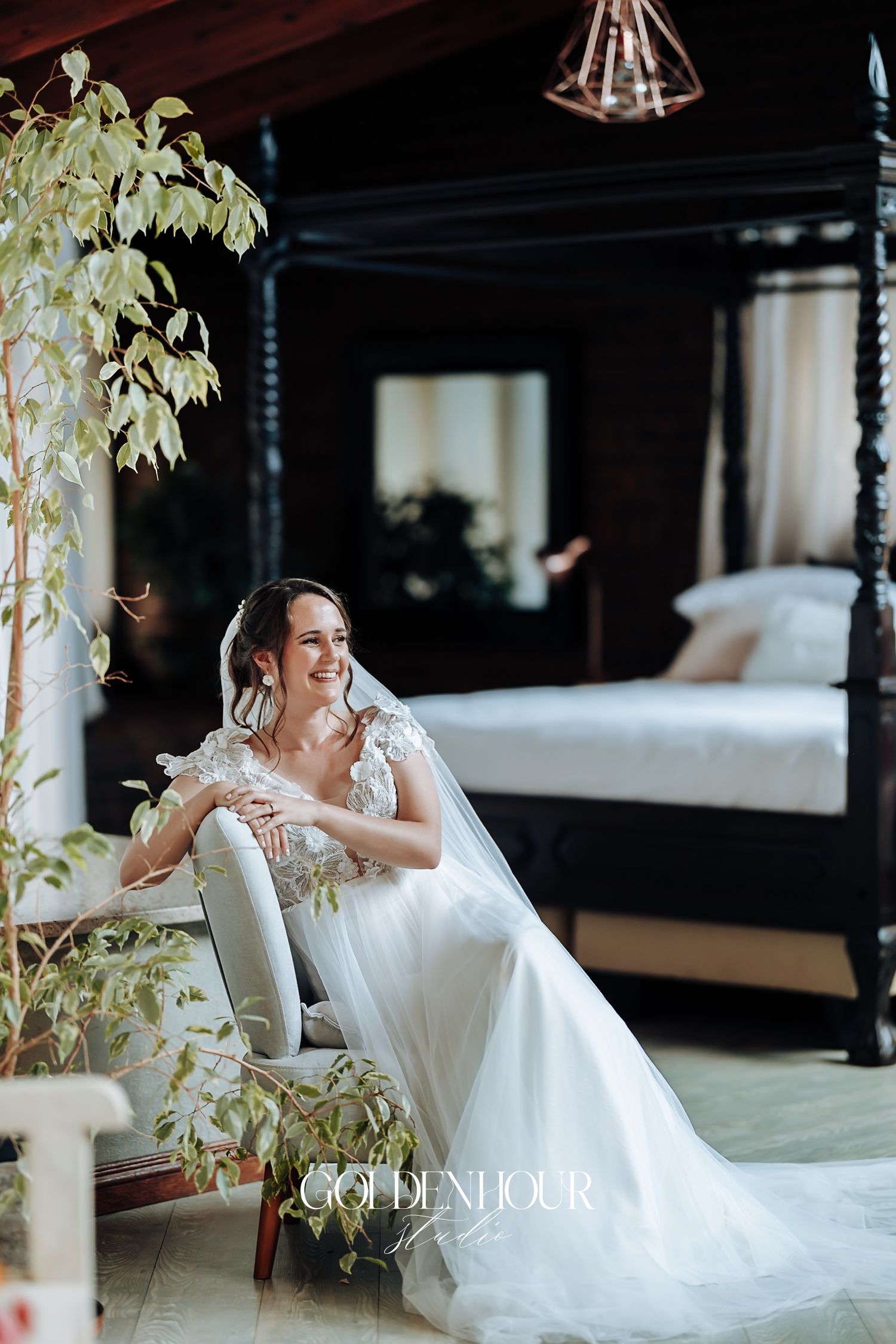 Radiant bride in lace wedding gown and veil seated in a chair inside an elegant dark-wood bedroom.