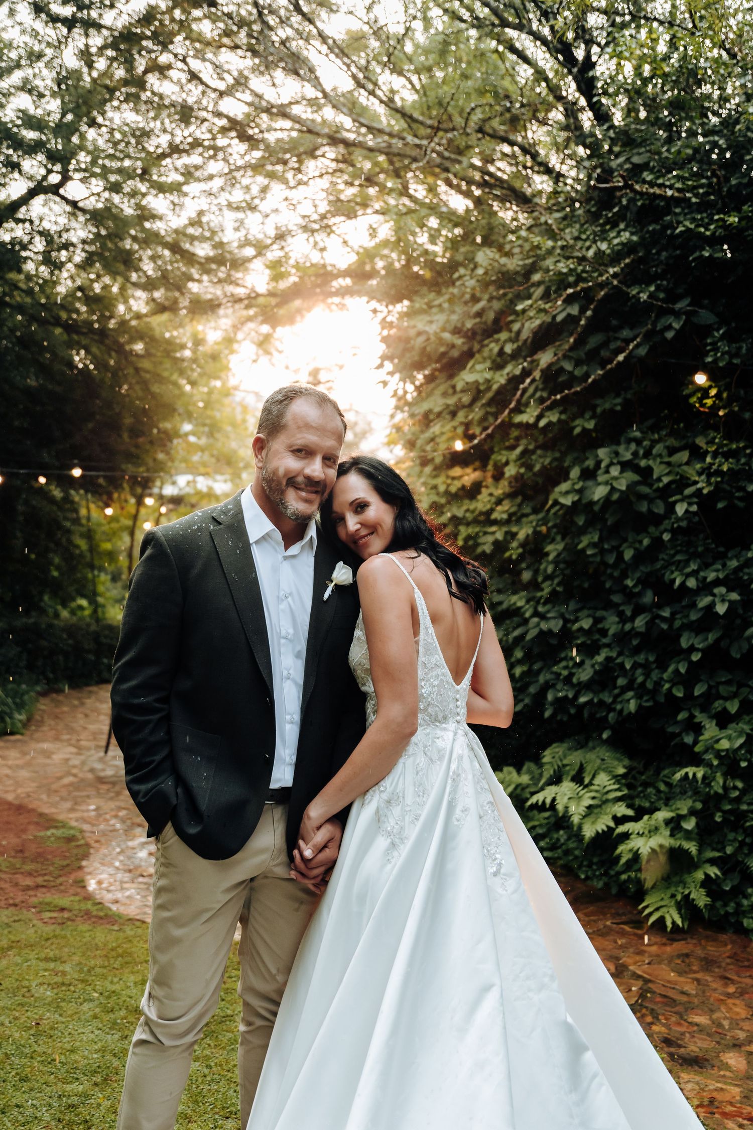 A smiling bride and groom hold hands in a lush garden at golden hour, she in a white gown, he in a dark blazer.