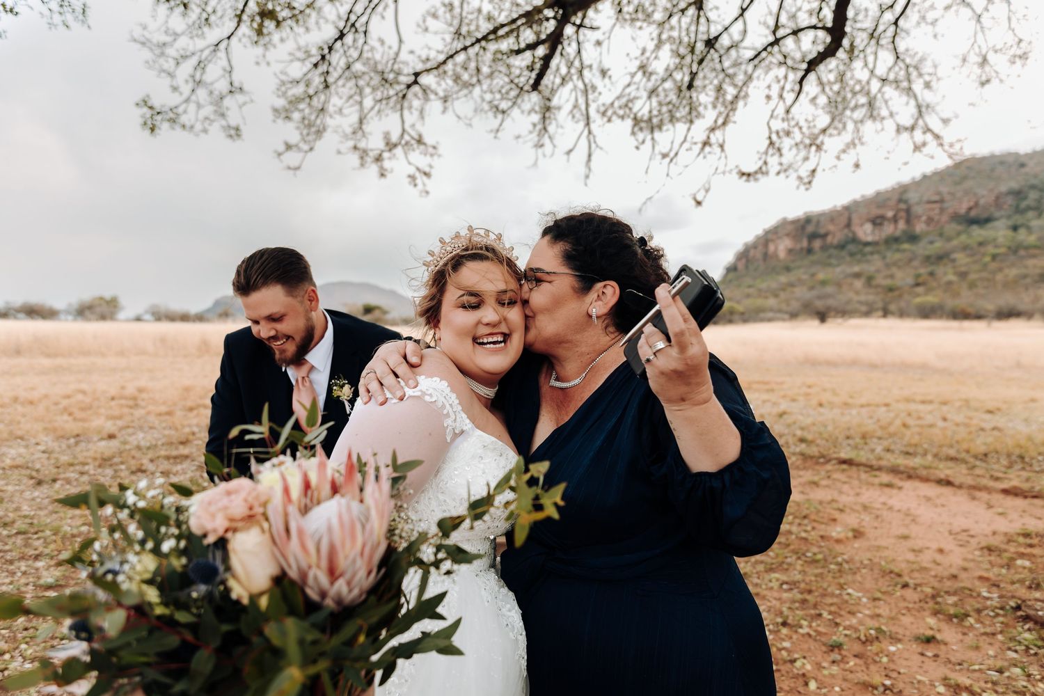 Three people laughing and embracing while holding floral bouquets in an outdoor desert landscape setting.