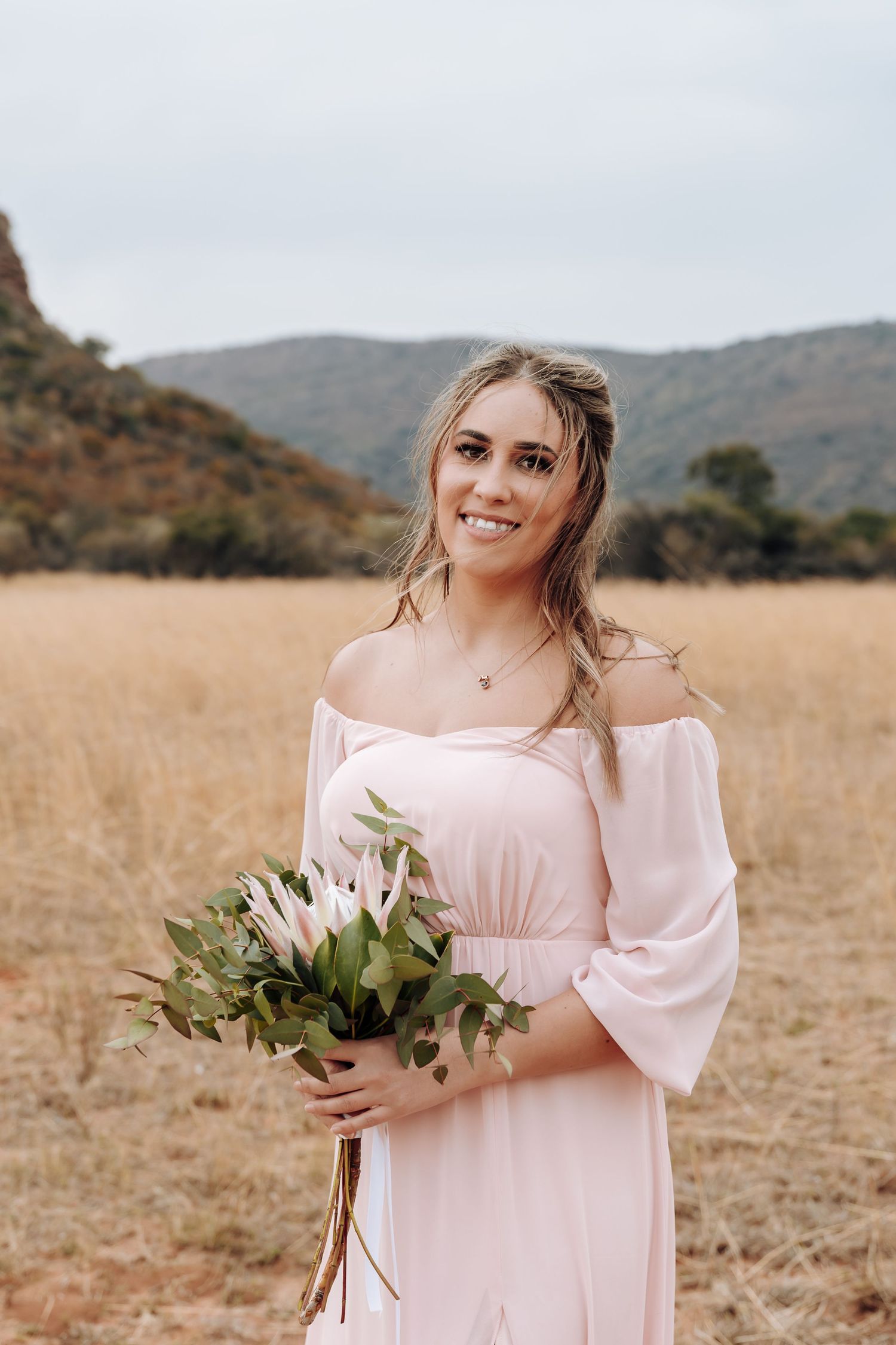 A person in a blush-pink off-shoulder dress stands in a golden field holding a bouquet of white lilies.